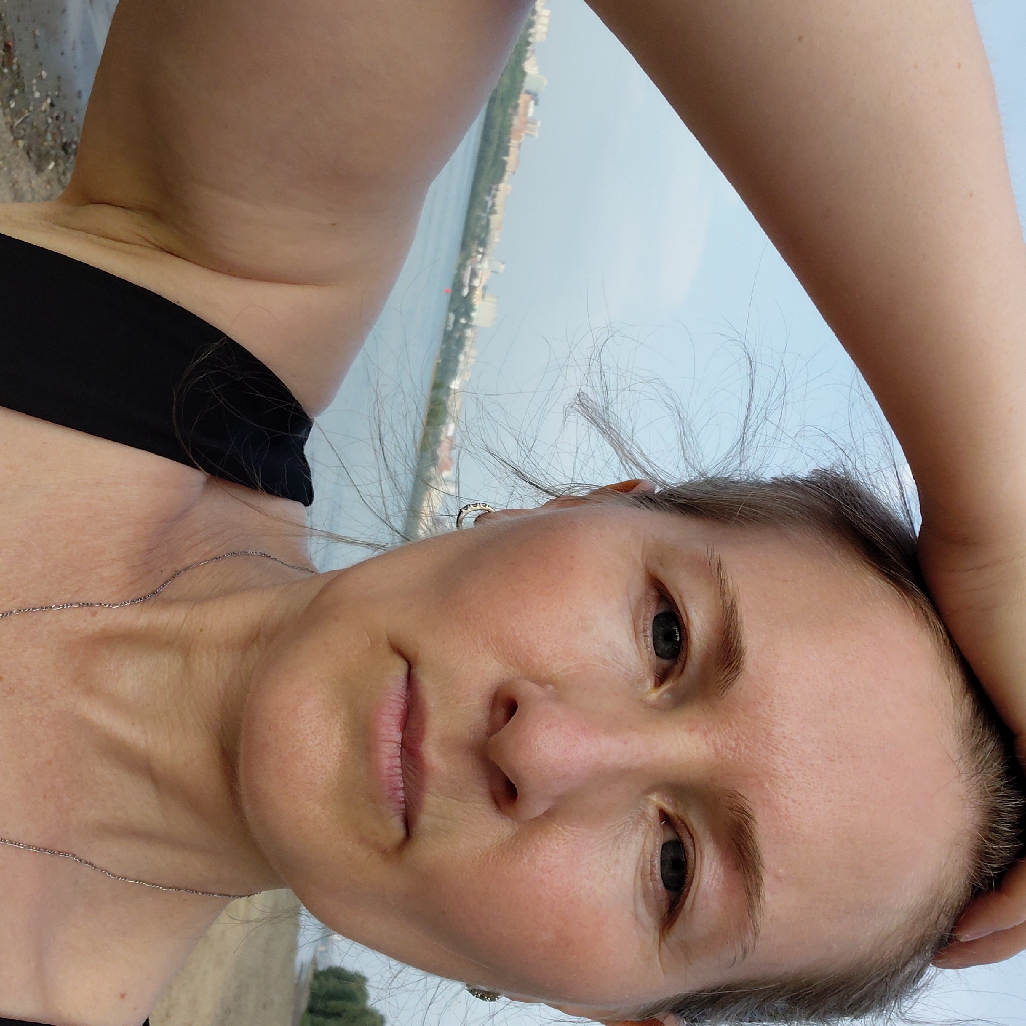 A woman with light skin, blonde hair, and blue eyes is taking a selfie outdoors. She is wearing a black top and a silver necklace. Her right arm is raised, touching the top of her head. The background shows a body of water, a sandy shore, and a city skyline with buildings and trees, possibly indicating the city of Novosibirsk, Russia, given the presence of the Ob River and its urban landscape. The sky is light blue and hazy, suggesting it may be a sunny or overcast day.