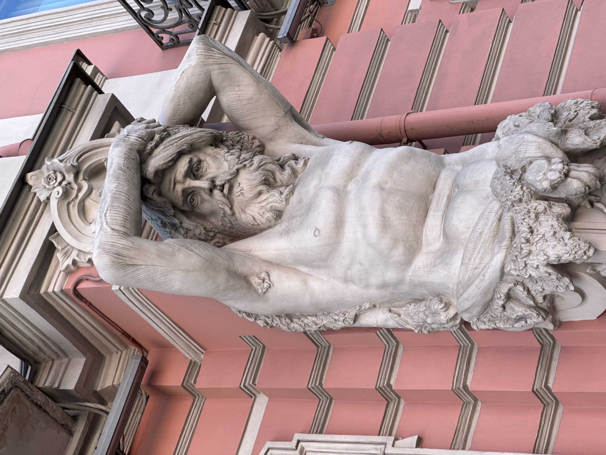 This image captures an upward-angled view of an architectural Atlas figure on the facade of the Singer House (Dom Knigi / House of Books) at Nevsky Prospekt, 28, Saint Petersburg, Russia. The central subject is a large, light grey stone or concrete statue of a muscular, bearded male figure. He is depicted with long, flowing hair and a solemn expression, his hands clasped over his head as if supporting a heavy structure above. His bare torso exhibits sculpted musculature, and his lower body is partially draped, transitioning into ornate, organic, foliage-like carvings at the base. The statue displays visible cracks, notably on the right arm.
The Atlas is set against a vibrant salmon-pink stucco building facade, characteristic of the Singer House. White or light grey classical architectural moldings and cornices horizontally segment and frame the pink walls, creating a tiered effect. Several utility elements are visible: a prominent rust-colored metal downpipe runs vertically along the right side of the statue, with additional rust-colored and darker electrical conduits adhering to the building surface. In the upper right corner, a section of a black, ornate wrought-iron balcony railing is present, along with a sliver of a sky-blue reflected window. Natural light illuminates the scene, highlighting the textures and details of the sculpture and architecture.