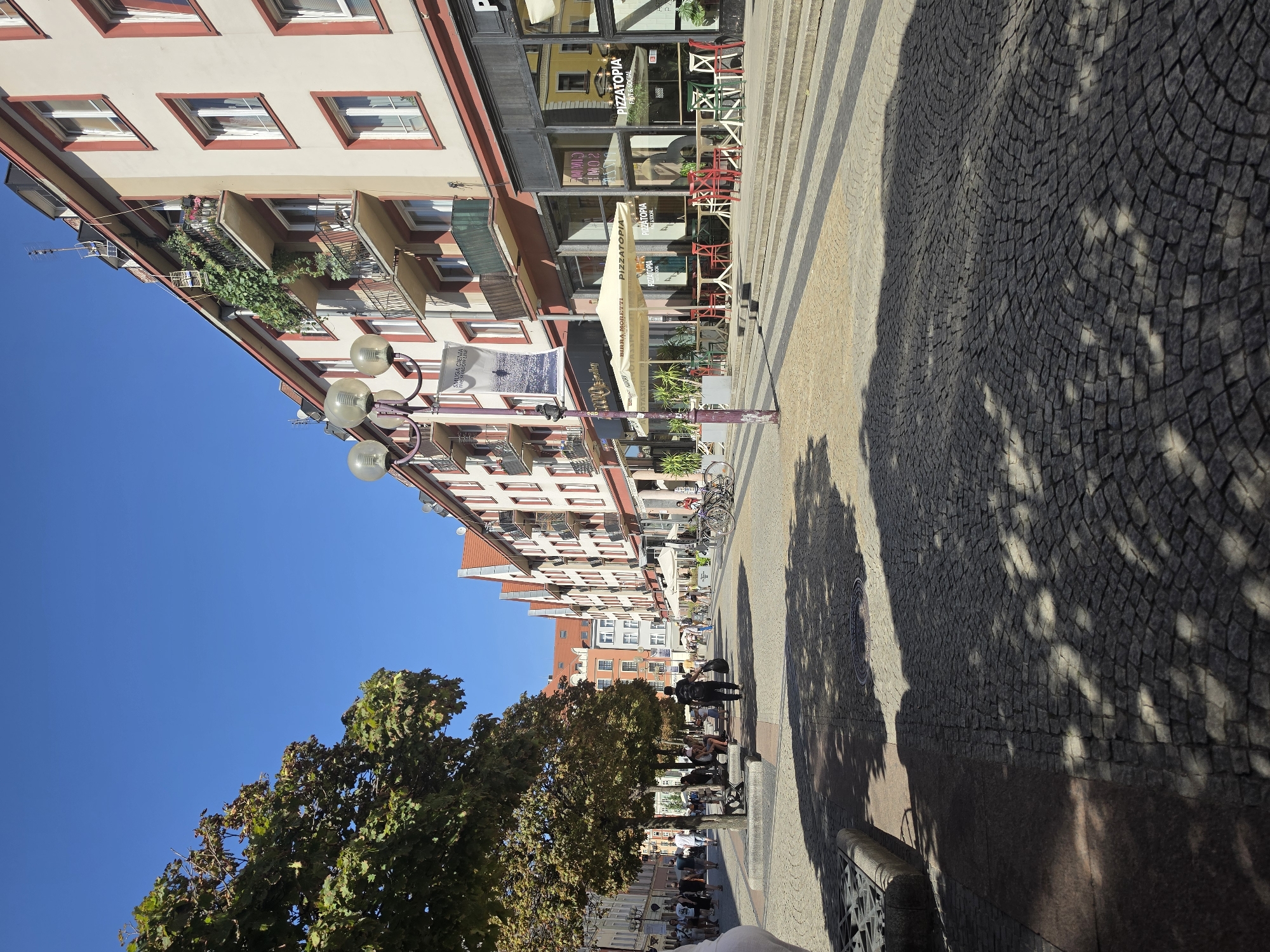 This is a street scene in Wrocław, Poland, likely in the city center. The image depicts a cobblestone pedestrian street lined with multi-story buildings. The buildings feature a mix of architectural styles, with some exhibiting balconies adorned with greenery and flowers. Several businesses, including "PIZZATOPIA Fast & Social," are visible on the ground floor, with outdoor seating areas under awnings. A lamppost with multiple spherical lights stands near the sidewalk. Trees with lush green foliage cast dappled shadows on the ground, indicating a sunny day. People are seen walking and sitting on benches, suggesting a leisurely atmosphere. Bicycles are parked near the establishments. The sky is a clear, vibrant blue.