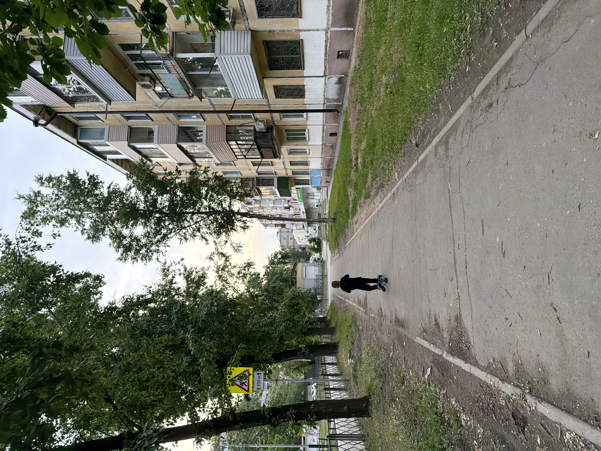 The image depicts a residential street scene in Khabarovsk, Russia, under an overcast sky. In the foreground, a child wearing black clothing is riding a scooter on a concrete sidewalk. The sidewalk is bordered by a strip of grass and then a line of mature trees on the left, and a multi-story apartment building on the right. The building is yellow and beige with numerous balconies, some enclosed and others open. A yellow triangular traffic sign with a black silhouette of children crossing and text indicating "300 M ↑" is visible on the left side of the sidewalk. The street appears to be a typical urban environment with a sidewalk, road, and residential buildings.