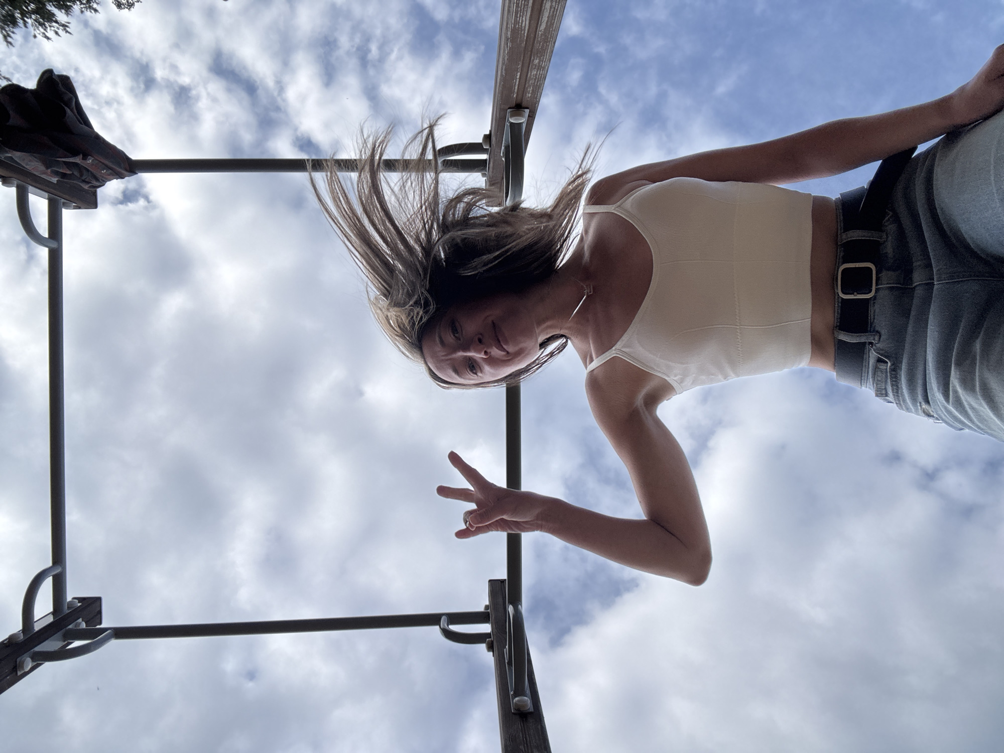 A low-angle shot features a person hanging upside down from playground equipment, likely in an outdoor public space. The person, a light-skinned female, is wearing a white tank top and denim jeans with a black belt. Her hair is cascading downwards, suggesting she is fully inverted. She is holding up a hand in a "peace" sign. The background is a cloudy sky. The playground equipment appears to be made of dark metal, and the wooden top of the pull up bar.
The location cannot be confirmed as Chaikovsky, Russia, without additional context or visual cues.