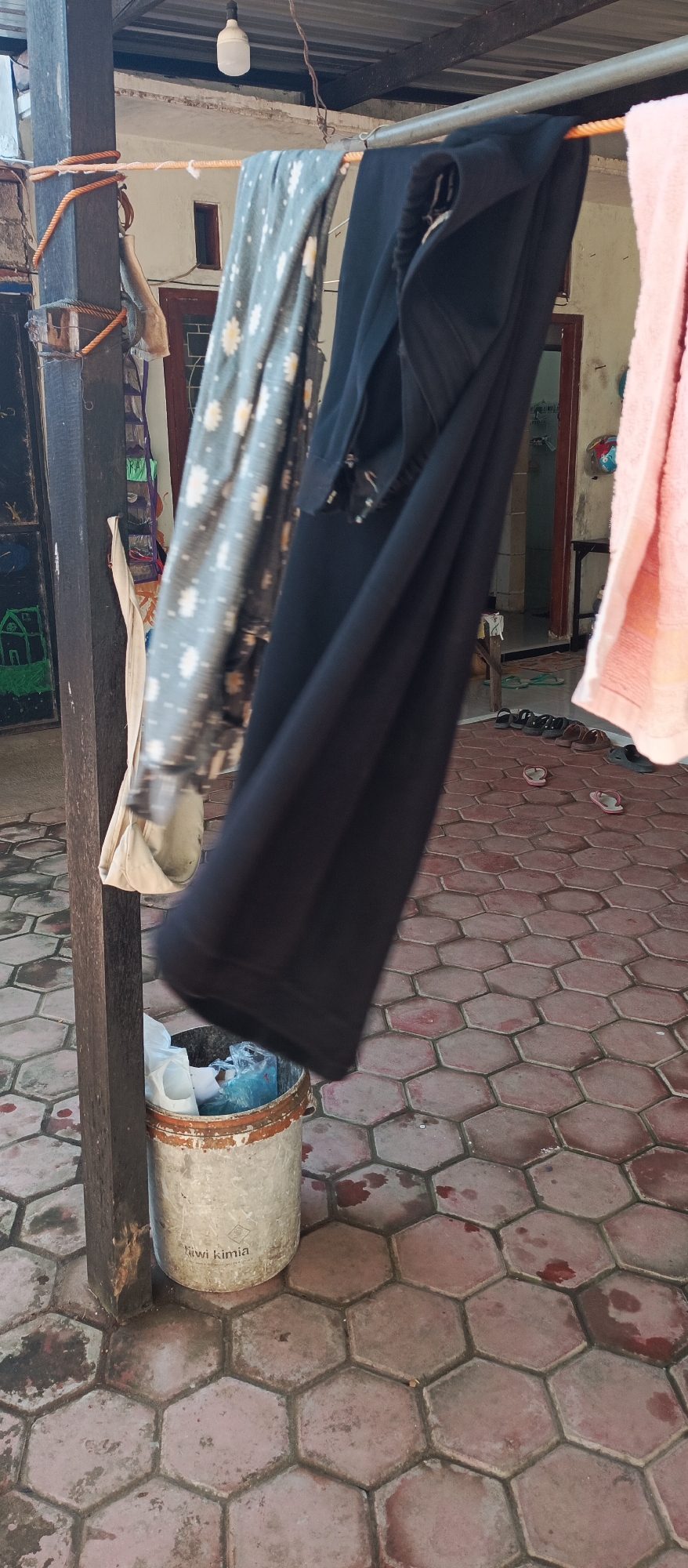 An outdoor scene in Jatisari, Indonesia, features laundry hanging on a clothesline above a tiled ground. A metal clothesline is suspended between a wooden post and a building structure. Several items of clothing are draped on the line, including a black pair of pants, a patterned grey fabric, and a pink towel. To the left of the clothesline, a wooden post is secured with orange rope. At the base of the post, a weathered metal bucket is filled with white plastic bags and other refuse. The ground is paved with hexagonal terracotta tiles, some of which are stained. In the background, a doorway leads into a building, and several pairs of flip-flops are scattered on the ground. The lighting suggests it is daytime.