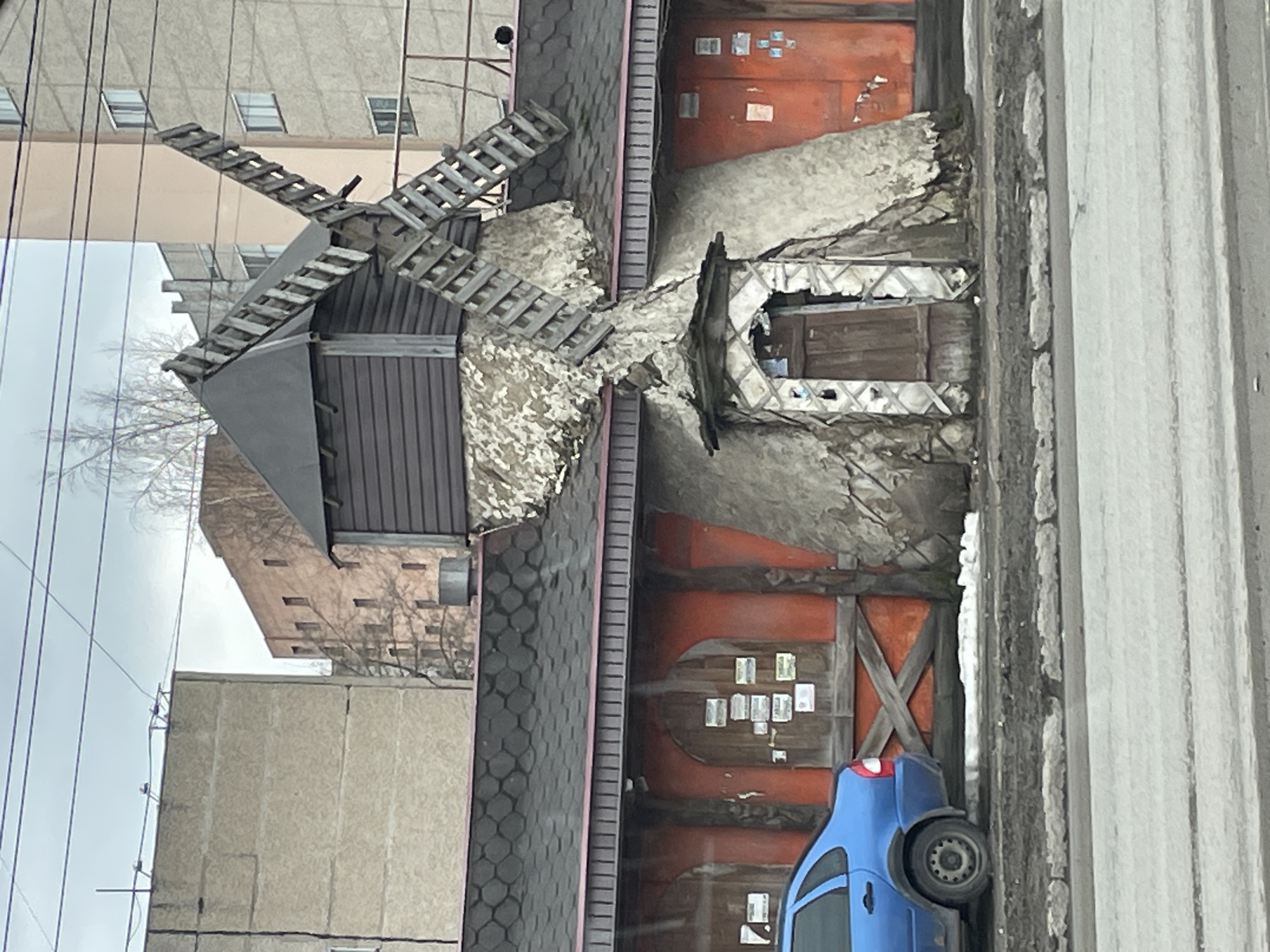 The image depicts a small, decorative windmill structure next to a building in Izhevsk, Russia. The windmill is composed of a dark wooden base with gray blades, and is partially covered with a light-colored, possibly deteriorating, material. Adjacent to the windmill is a dilapidated building or wall structure with an arched entryway and exposed timber framework, painted in rusty orange. A blue car is parked near the building. The surrounding environment includes a multi-story apartment building in the background and a gray corrugated metal structure on the right side of the image. The overall scene suggests an urban setting with some degree of disrepair.