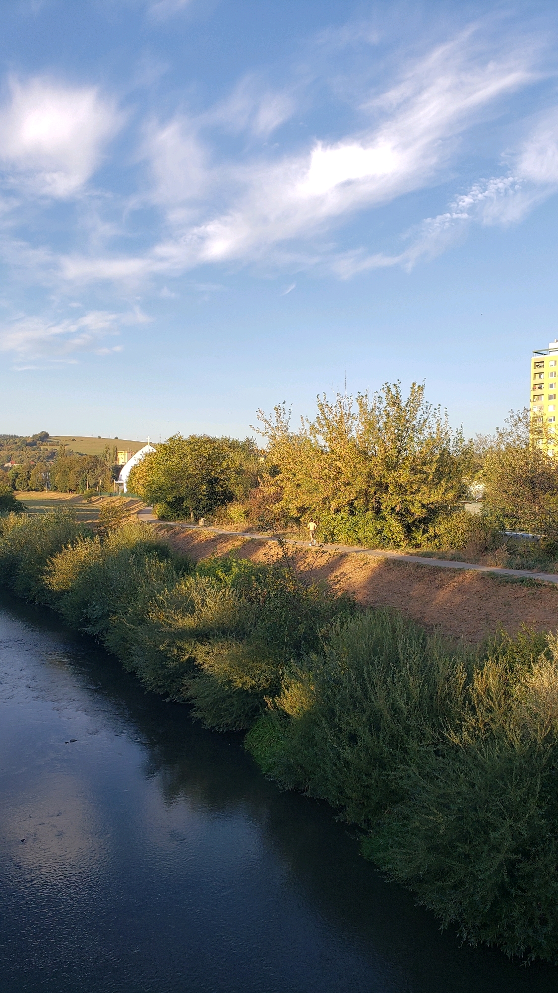 The image captures a scenic view of the Torysa River in Prešov, Slovakia, during what appears to be late afternoon or early evening. The river flows from the bottom left towards the top right of the frame, its dark, reflective surface broken by subtle ripples. Along the riverbank, dense green foliage lines the water's edge, with taller trees and bushes situated on a higher embankment behind them. A dirt path runs along this embankment, and on it, a lone individual, appearing to be a male in light-colored clothing, is jogging towards the right side of the image.
The background reveals a park-like setting with more trees, some displaying autumnal colors. Further in the distance, a distinctive building with a white facade and a prominent cross on its roof is visible, likely a church or religious institution. To the far right, a tall, yellow apartment building stands, indicating the proximity of urban development to this natural area. The sky above is a vibrant blue, sparsely populated with wispy white clouds, suggesting fair weather. The overall impression is one of a tranquil, well-maintained urban green space.