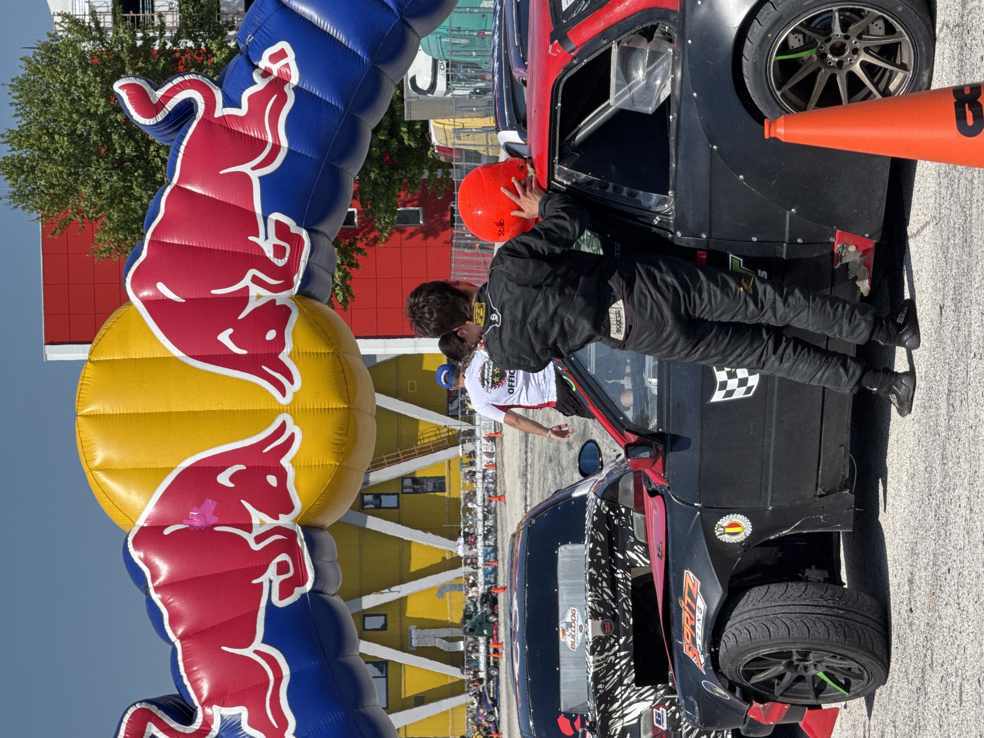 A man in a black racing suit and orange helmet stands next to a black race car with red accents under a blue, red, and yellow Red Bull inflatable arch. Another man is in the background wearing a white shirt with "Official" printed on it. Behind them is a multi-level yellow building with white diagonal supports. There are spectators and other cars in the background. The location is likely a racing event in Plovdiv, Bulgaria, given the presence of the Red Bull inflatable arch and the race cars.