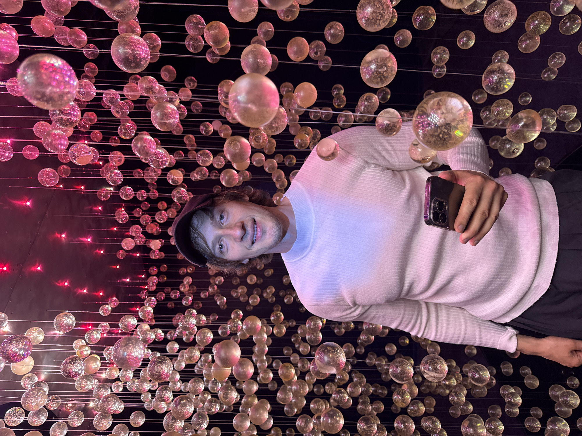 A man in a white sweater and a dark baseball cap is standing in a dark room filled with hanging glass spheres. The spheres are illuminated with pink and red lights, creating a glowing effect. The man is holding a smartphone in his right hand and looking at the camera with a smile. He is positioned in the center of the frame, with the spheres surrounding him on all sides. The background is dark, making the illuminated spheres stand out. It is not possible to determine the specific location in the city of Saint Petersburg, Russia from the image.