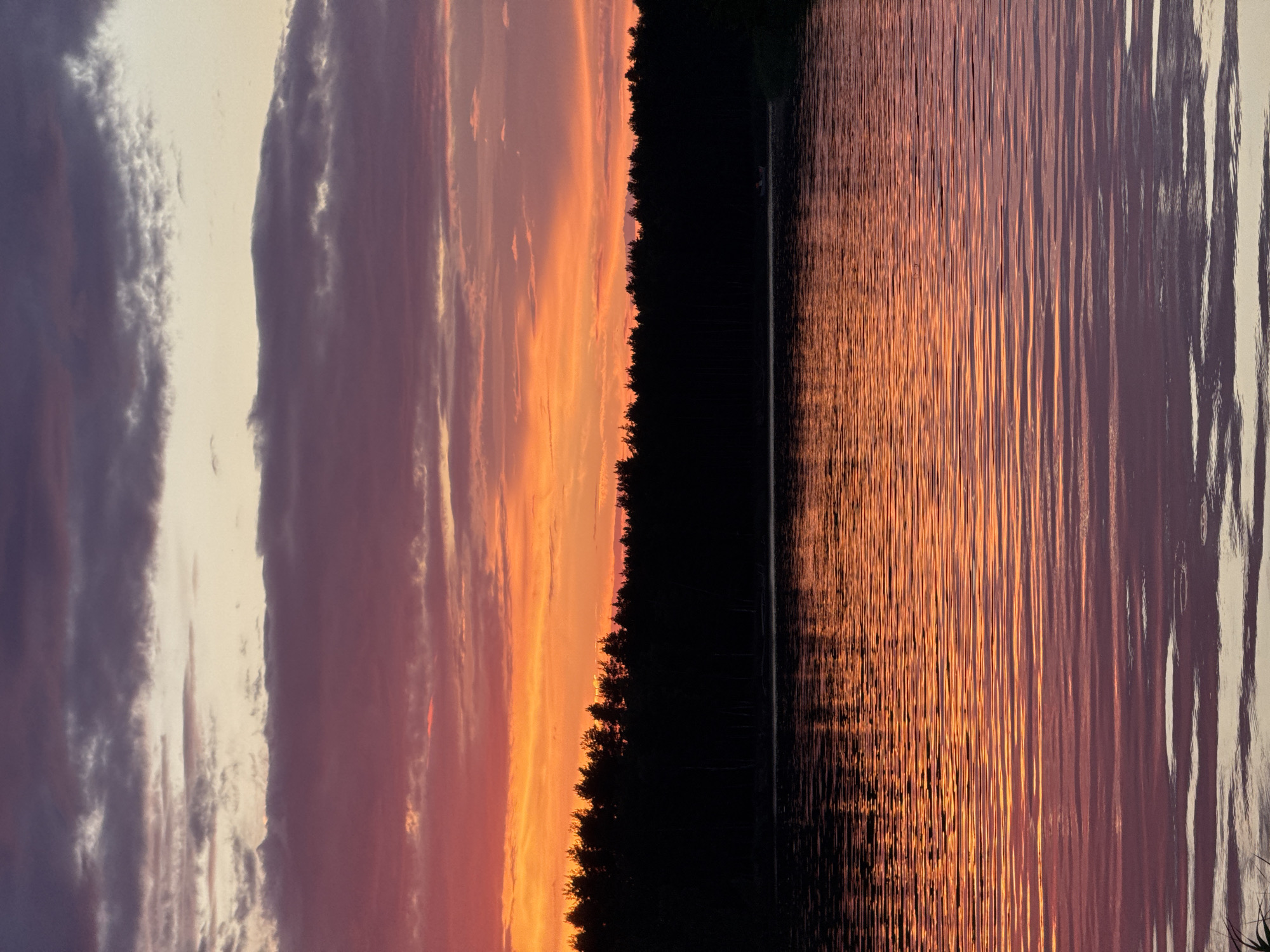 The image depicts a serene sunset over a body of water, likely a river or lake, in Carnikava, Latvia. The sky is ablaze with vibrant colors, transitioning from a soft yellow and orange near the horizon to deeper shades of pink and purple higher up. Clouds are scattered across the sky, catching the light and adding depth to the scene.
Silhouetted against the colorful sky is a dense line of trees, forming a dark, unbroken horizon. The water in the foreground reflects the dramatic colors of the sky, creating a shimmering, undulating surface. The reflections are broken into horizontal lines by the ripples on the water.
A small, light-colored boat is visible in the distance, positioned on the water near the tree line, suggesting a tranquil, outdoor recreational setting. The overall atmosphere is one of peace and natural beauty, characteristic of a Latvian sunset over its waterways.