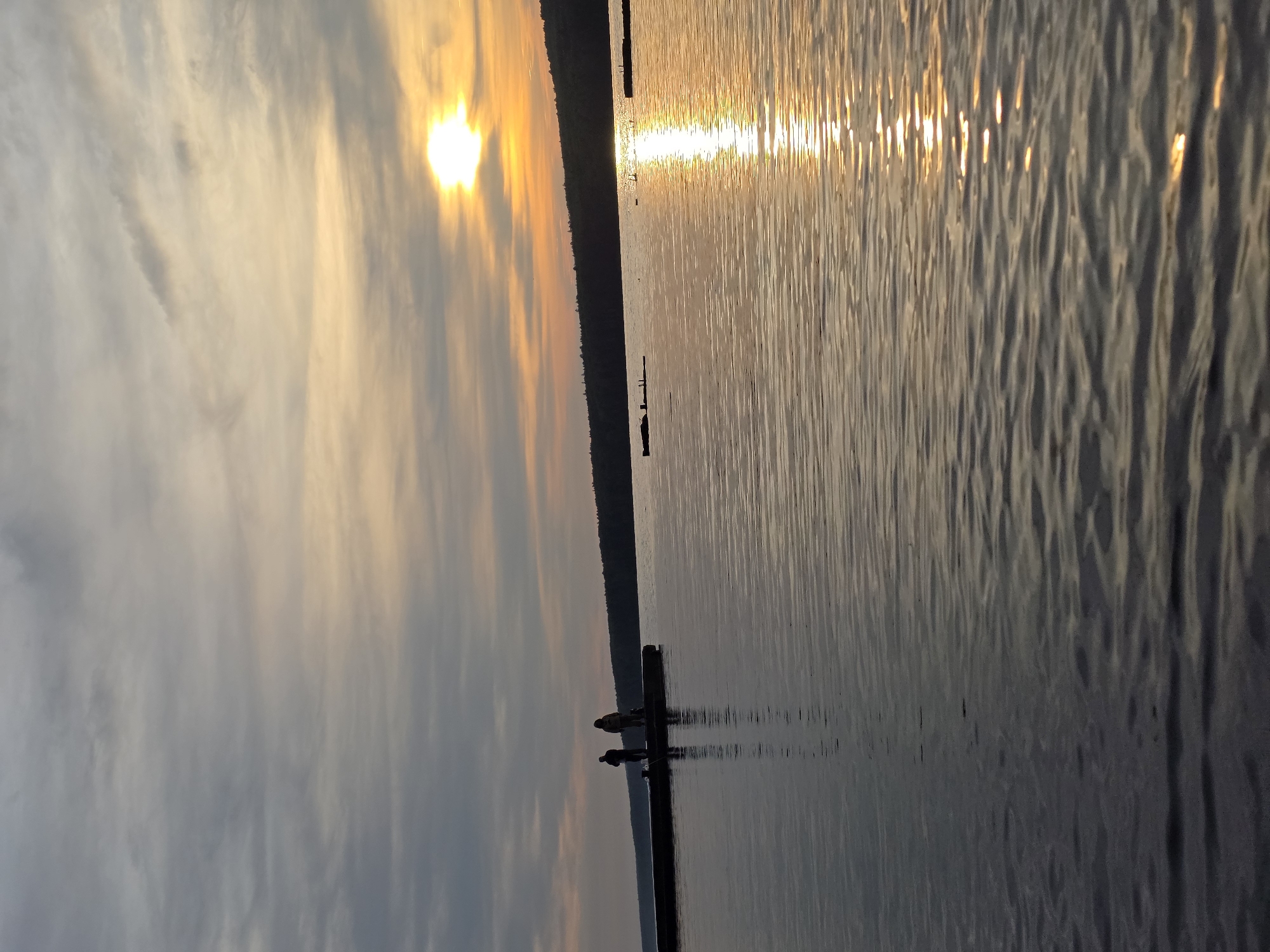 The image depicts a serene sunset over a wide body of water, characteristic of a large river or lake in Chaikovsky, Russia. The sun, a brilliant orb of yellow and orange, is positioned low in the sky towards the left, casting a strong glare and a shimmering path of light across the water's surface.
The sky above the horizon is adorned with soft, wispy clouds, transitioning from muted grey and white tones to warm golden hues closer to the sun. A dark, narrow landmass, likely a distant forested shoreline, spans horizontally, separating the sky from the expansive water.
In the midground, a long, dark pier or dock extends into the calm water from the right side of the frame. Two silhouetted figures stand on this pier, their forms sharply outlined against the illuminated water, suggesting they are observing the tranquil scene or possibly fishing. Their reflections are clearly visible in the water directly beneath the pier. The water's surface is gently rippled, catching and dispersing the sun's light in sparkling lines. Several other smaller, dark, vertical posts or structures are sparsely visible further out in the water. The overall impression is one of peacefulness at dusk. No visible text is present in the image.