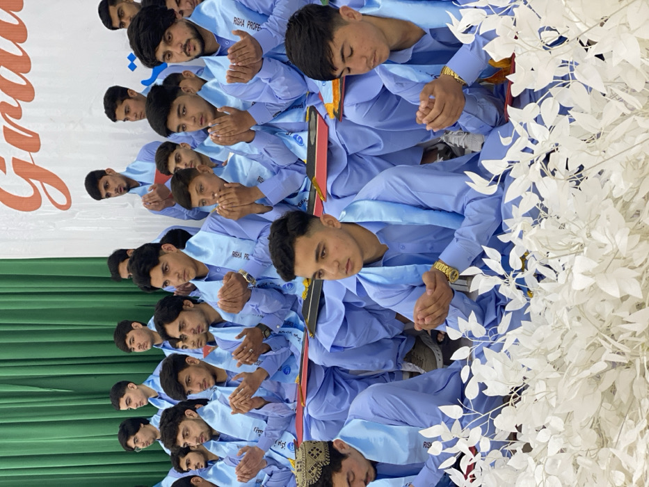 This image captures an indoor graduation ceremony in Kandahar, Afghanistan, featuring approximately two dozen young men. The individuals, primarily in their late teens to early twenties, are seated in rows, uniformly dressed in light blue long-sleeved shirts and matching trousers. Each wears a light blue stole over their shoulders, with some stoles bearing the text "RISHA PROF..." or "ریشا پرو..." (Risha Professional). Many participants have their hands raised with palms open and facing upwards, a gesture of prayer or supplication. Several individuals have black folders or small documents, some with red or yellow accents, on their laps. The background features a green curtain, partially covered by a white banner displaying the word "Gradu" in large orange letters, indicating "Graduation," alongside other Farsi/Dari text. In the lower left foreground, a participant wears a brown traditional Afghan cap, while another in the center foreground wears a gold-colored wristwatch. White artificial foliage decorates the immediate foreground, partially obscuring the lower bodies of some participants.