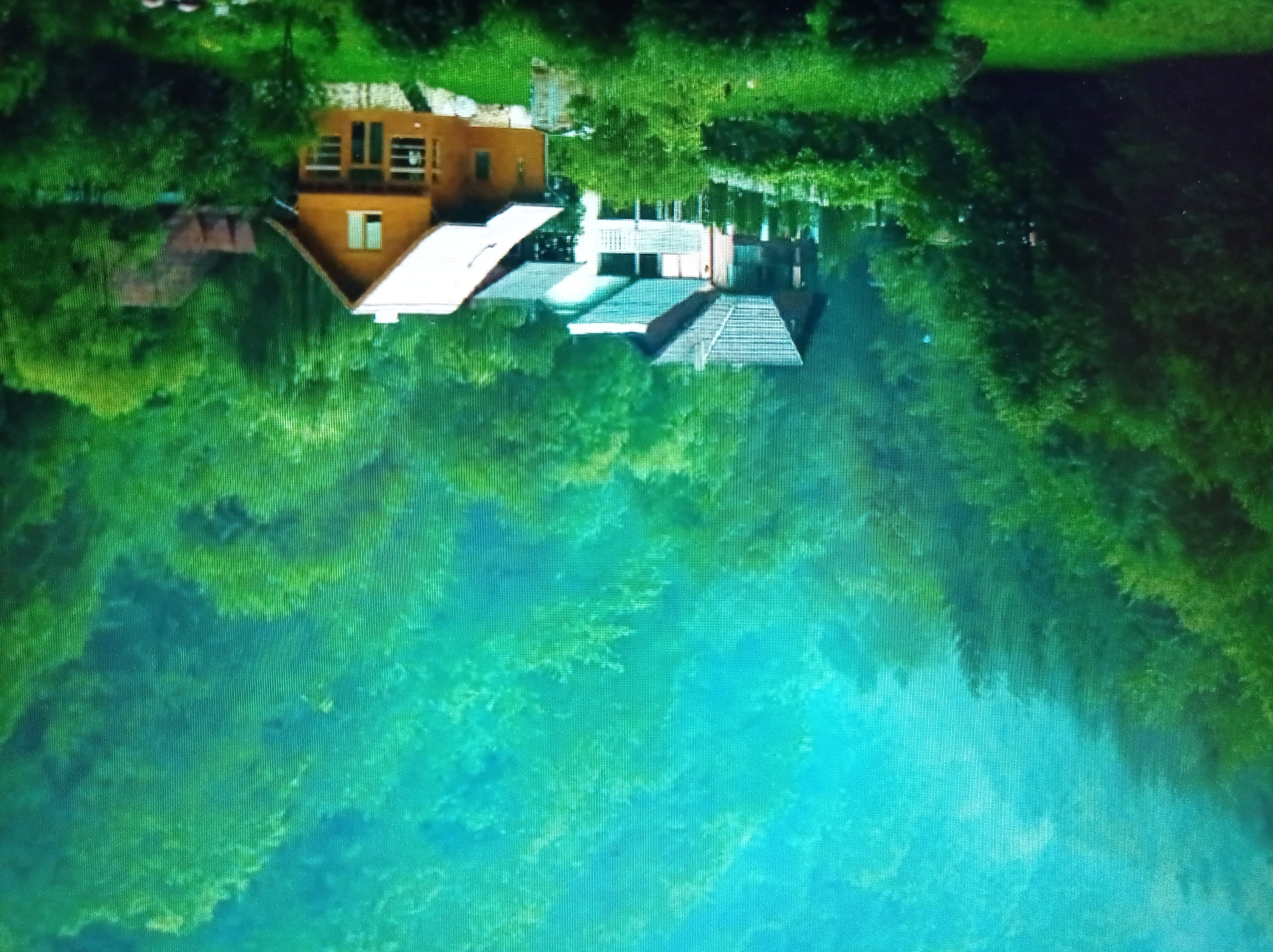 The image depicts a rural, mountainous landscape with several houses nestled amongst lush green trees. The sky above is a hazy, bright turquoise, possibly indicating mist or fog. In the foreground, a well-maintained grassy area slopes down. Several houses are visible: a prominent wooden house with a white roof and large windows, to its left a white house with a light blue roof, and further left, a darker house with a grey roof. The houses appear to be residential properties. The surrounding environment is dense with foliage, suggesting a forest or heavily wooded area. The overall atmosphere is tranquil and scenic. This image does not contain any elements that place it specifically in the city of Mukachevo, Ukraine, or any other identifiable urban location.
