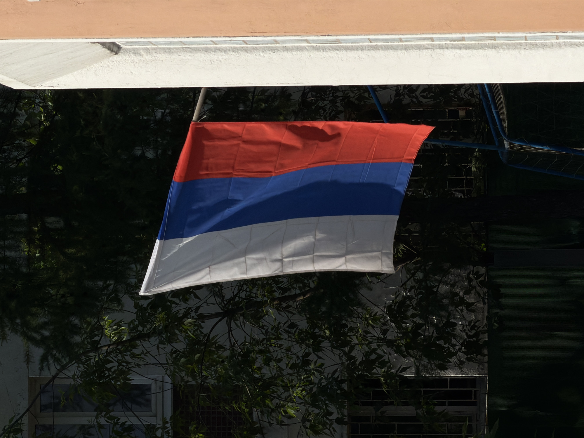 A flag with red, blue, and white horizontal stripes hangs from a pole attached to a building. The flag is slightly wrinkled, and a hint of sunlight illuminates the white stripe. The building above the flag is painted with two tones of beige. Behind the flag are tree branches with green leaves, and a building with a window. A blue metal structure is visible to the right of the flag. The exact location in Moscow cannot be determined from the image alone.