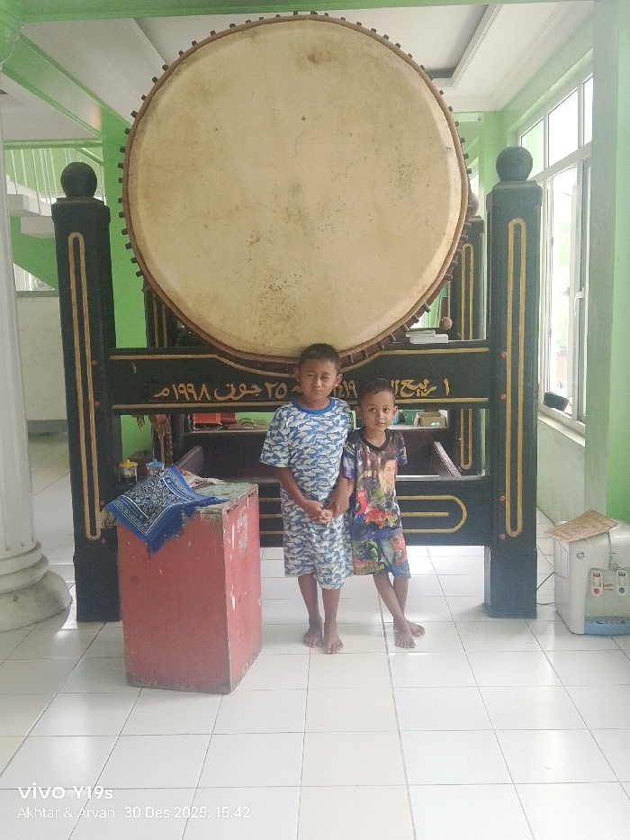 An indoor scene in Jagapura Kidul, Indonesia, features two young males standing barefoot in front of a large, circular, light-colored drum mounted on a dark, ornate wooden stand. The drum's surface shows signs of age and discoloration. The stand is black with gold decorative elements and displays Arabic script, including the visible date "٢٥ جون ١٩٩٨" (June 25, 1998). The taller child on the left wears a blue and white camouflage-patterned shirt and shorts, with his hands clasped. The shorter child on the right wears a colorful patterned shirt and shorts; his left hand is held by the taller child's right hand. Both children look directly at the camera with neutral expressions. To the left of the children, a reddish-brown rectangular wooden box holds a folded blue prayer mat with gold embroidery. To the far right, a white water dispenser with a brown top is positioned near large windows. The environment consists of light green painted walls, a white tiled floor, and a white pillar on the left. White stairs are visible in the upper-left background.