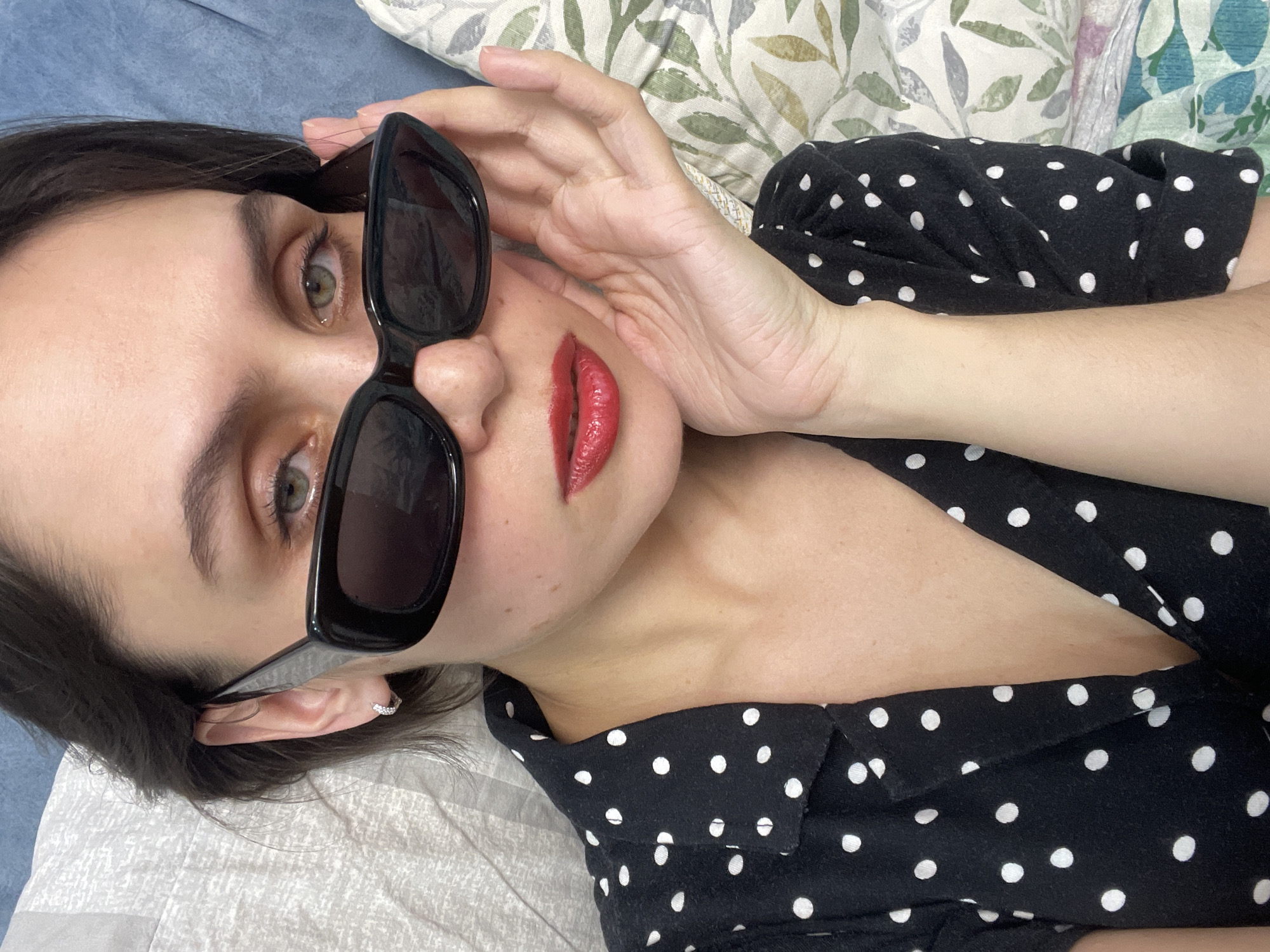 A woman lies on her back, partially covered by a bedsheet and pillow with a floral pattern. She wears a black dress with white polka dots and holds sunglasses up to her face with her right hand. She has light skin, green eyes, and dark hair. Her lips are painted red. There is no indication of Izhevsk, Russia.