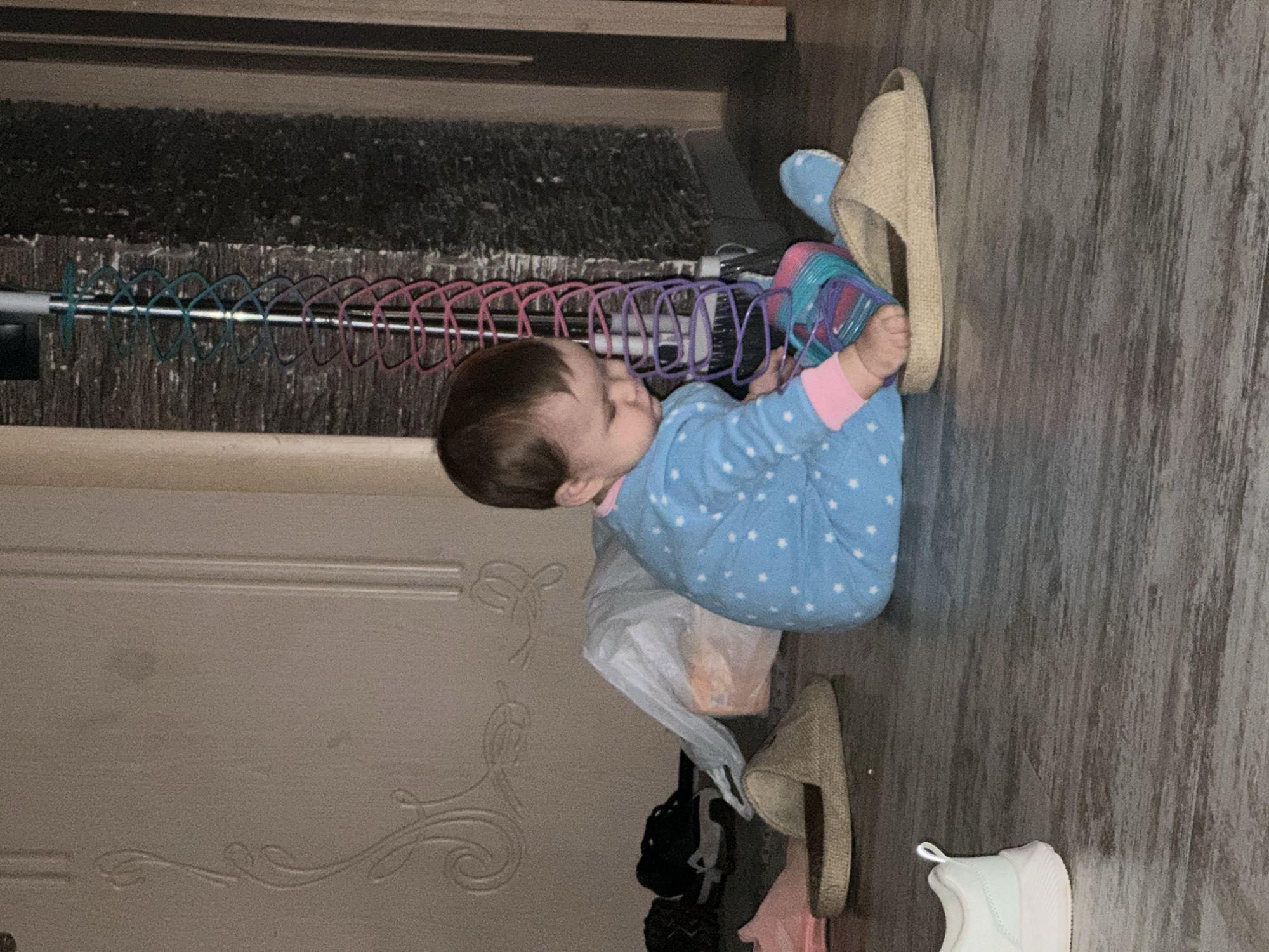 A toddler in blue pajamas with white stars sits on a grey wooden floor. The toddler is reaching into a sparkly rainbow slinky toy that is partially inside a tan slipper. A white sneaker is to the left of the toddler. A white plastic bag is behind the toddler, and black and grey shoes are to the left of the bag. A wall with a decorative design is behind the toddler. A tall, thin rack with colorful loops is to the right of the toddler. The scene appears to be indoors.