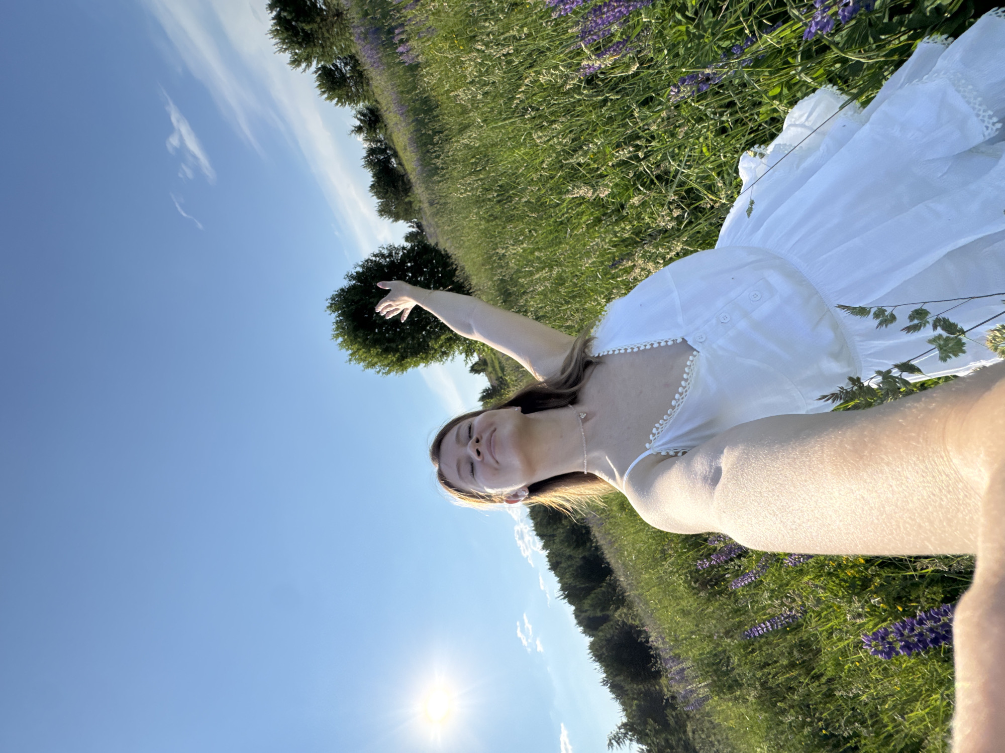 A young woman stands in a field of tall green grass and purple flowers under a bright blue sky with scattered clouds. She is wearing a white sundress and has her right arm raised. The sun is shining brightly in the upper left corner of the image. The background features a line of trees. This scene is located in the village of Van'ki, Russia.