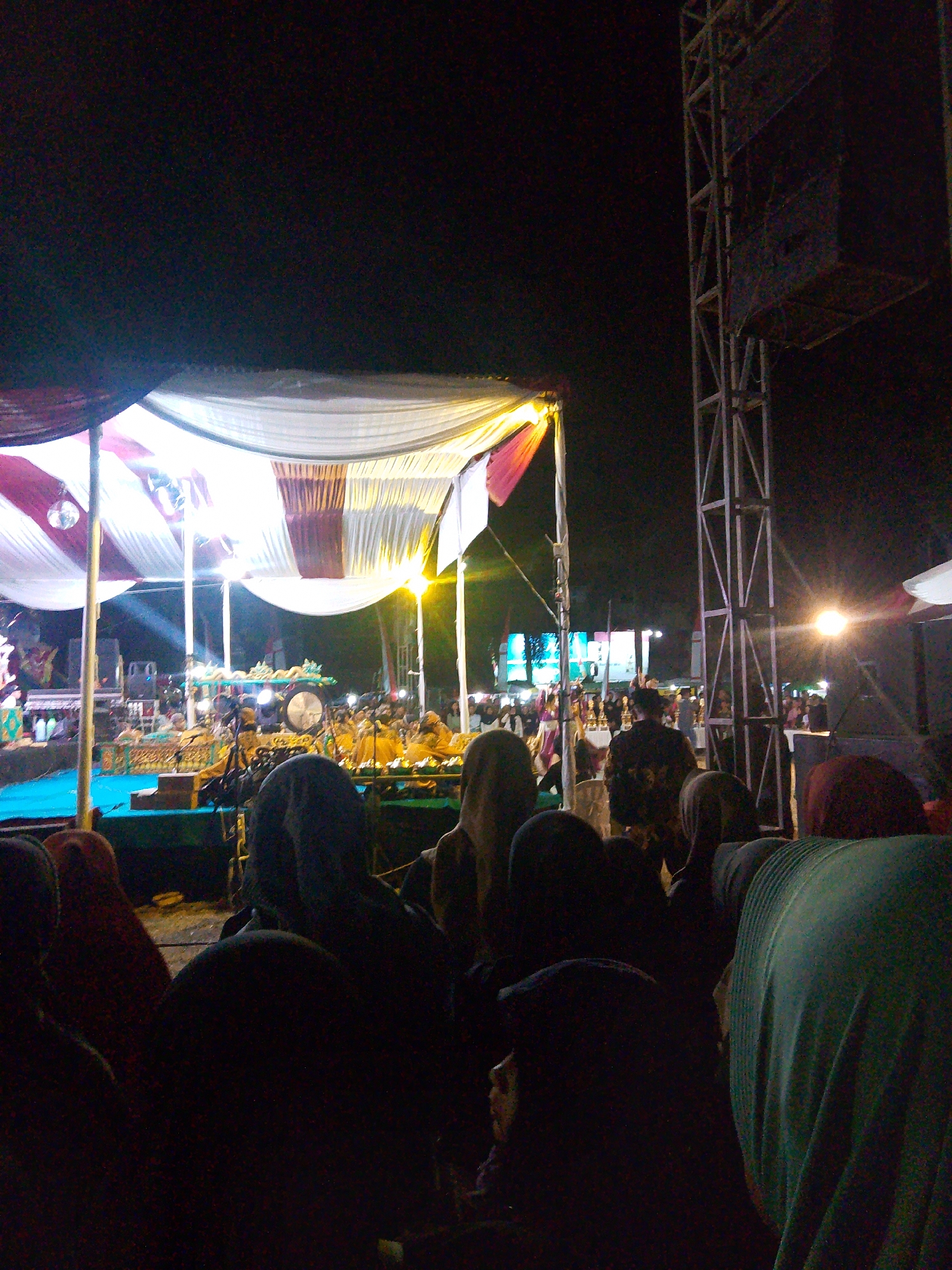 This image captures an outdoor evening event, possibly a cultural performance or festival, in Wanareja, Indonesia. In the foreground, a crowd of spectators is visible, primarily depicted from the back and shoulders up. Many are wearing headscarves or hijabs in various colors, including dark blue, red, and green. The lighting is dim, suggesting it's nighttime, with some artificial lights illuminating the scene.
Behind the crowd, a stage is set up under a draped canopy of white and maroon fabric. The stage is crowded with performers and musical instruments, including what appears to be a large gong or drum. There are also speakers and other technical equipment visible. A tall, metal scaffolding structure stands to the right of the stage, likely supporting lights or sound equipment. Bright lights are focused on the stage, creating a dramatic effect. In the background, more people are gathered, and there are some illuminated structures, possibly buildings or additional stages. The overall atmosphere seems to be one of communal gathering and entertainment.