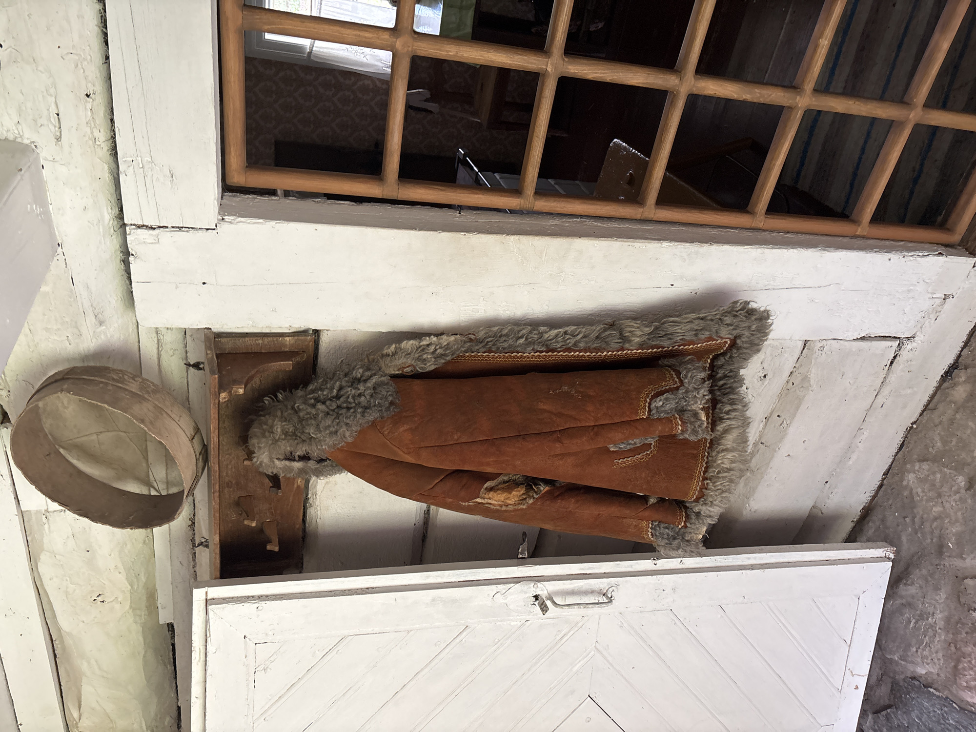 This image shows an interior scene within a rustic, log-built structure in Sielachowskie, Poland. A heavy, brown, fur-lined coat is hanging on a wooden rack mounted on a white-painted wooden wall. To the left, a white-painted wooden door with diagonal paneling is ajar. To the right, a wooden lattice window frames a view into another room, where a patterned wallpaper and furniture are visible. An old wooden sieve or colander is resting on a beam above the coat. The floor appears to be made of stone or concrete. The overall impression is one of an old, well-worn dwelling, possibly a historical or preserved building.