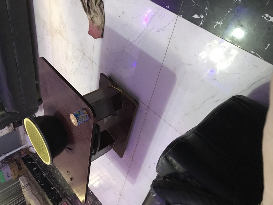A table with a can and a black object is positioned on a tiled floor with white and black sections. A dirty rag lies nearby. To the left of the table is a black object, possibly a chair. A dark-colored chair or similar object appears to the right. The room is illuminated with an artificial light source causing reflections on the floor.
