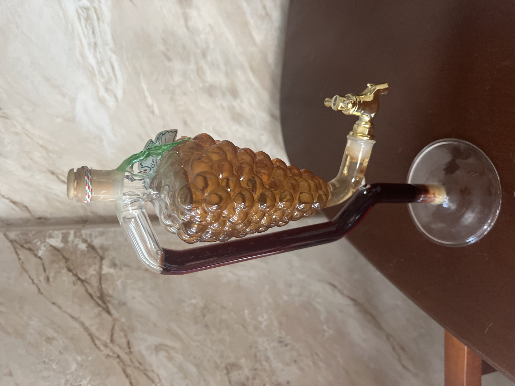 A novelty glass decanter shaped like a bunch of grapes, filled with amber-colored liquid, stands on a dark wooden surface. The decanter is supported by a glass stem and base, and has a small gold-colored spigot attached to its side. A red liquid is visible in a glass tube that runs from the top of the decanter down to the spigot. The background features a wall with a marble-like pattern. There are no people visible in the image. The location in the city of Vladimirovka, Russia is not discernible from the image.
