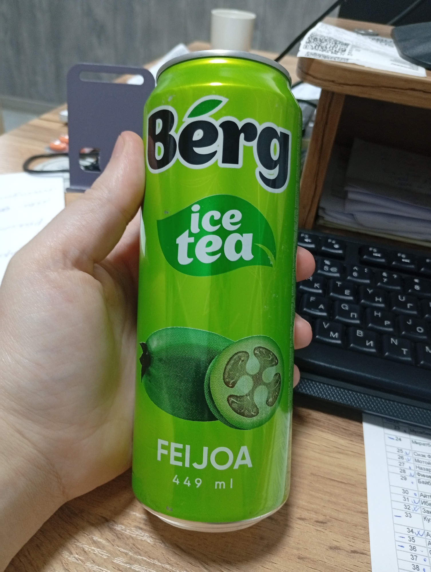 In the city of Osh City, Kyrgyzstan, a can of Berg Ice Tea Feijoa flavor, 449 ml, is held by a person's hand. The can is bright green and features the brand name "Berg" in large black letters, with "ice tea" written below in white text. An image of a feijoa fruit, halved to show its cross-section, is visible on the lower portion of the can. The background shows a wooden desk with a keyboard, a stack of papers with writing on them, and a grey office organizer.