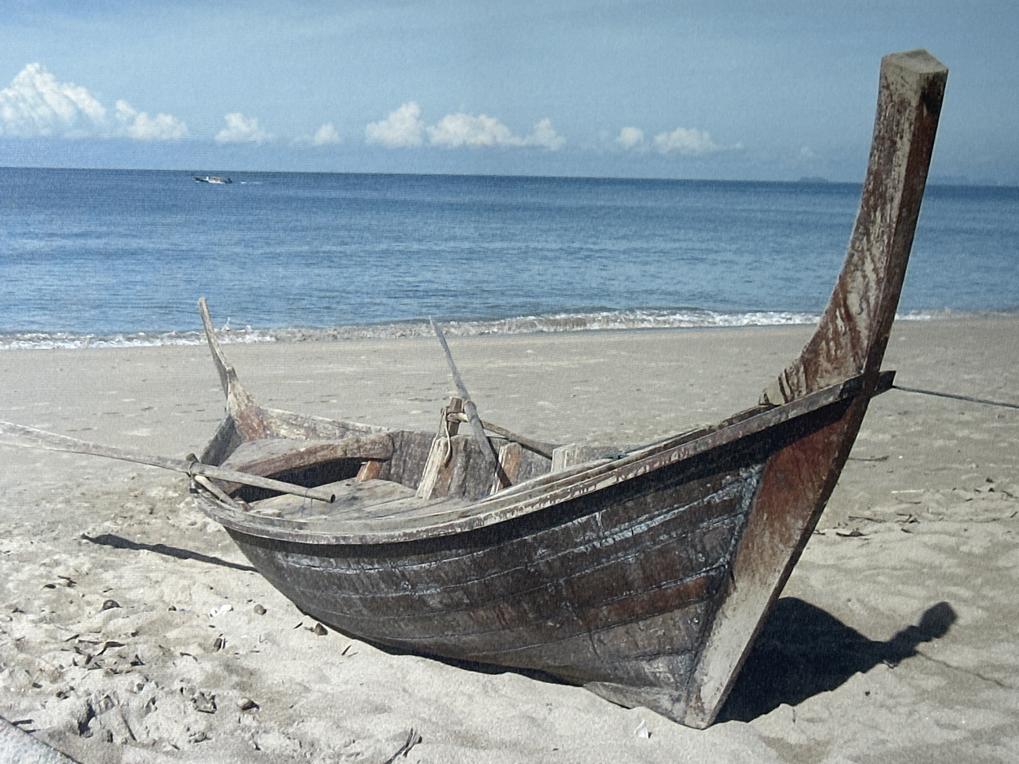 A weathered, wooden fishing boat is beached on a sandy shore. The boat is dark brown and appears old, with visible planks and signs of wear and tear. A long, wooden oar is propped against the side of the boat, extending outwards. The boat is angled, with its bow pointing towards the right of the frame and its stern towards the left.
The setting is a beach with a gentle slope leading down to the ocean. The sand is a light tan color and appears dry, with some scattered debris. The ocean is a calm, deep blue, with small waves lapping at the shore. In the distance, the horizon is a clear line between the sea and the sky. The sky is a pale blue, with a few scattered white clouds. There is another, smaller boat visible in the far distance on the water, which appears to be moving. The overall impression is one of a quiet, possibly isolated, coastal scene. The lighting suggests it is daytime, likely late morning or early afternoon, with a bright sun casting shadows. There is no visible text in the image.