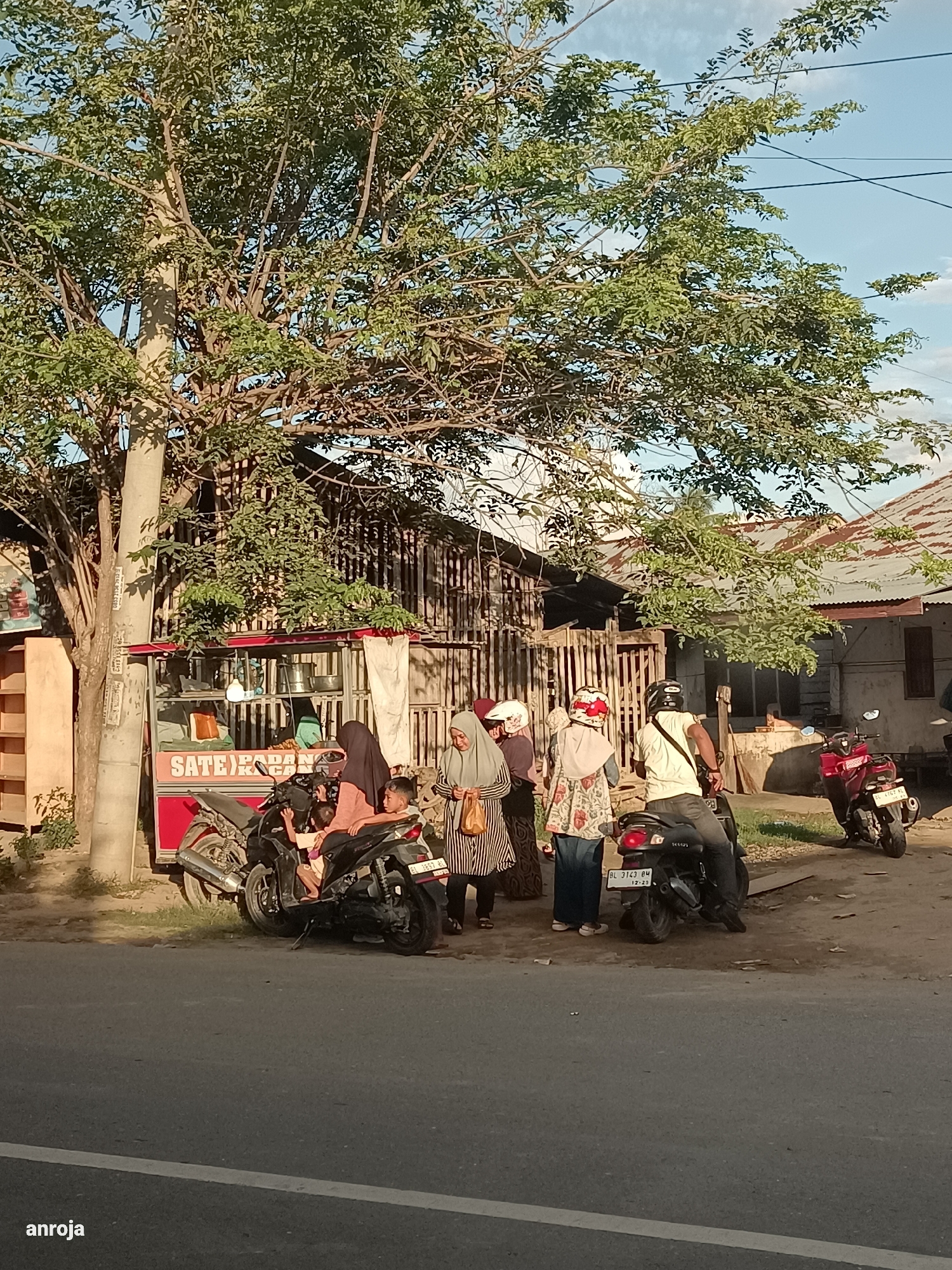 This media file depicts a street scene in Blang Cut, Indonesia, likely during the late afternoon or early evening, given the warm, golden light and long shadows. The primary subjects are a group of people gathered around a small food stall selling "Sate" and "Panga". Several individuals, including women wearing hijabs and a young boy, are interacting near the stall and two motorcycles. One motorcycle is parked in front of the stall with a woman and child on it, while another is parked further down the street. A man wearing a helmet is seated on a third motorcycle.
The scene is set alongside a paved road, with a large tree providing shade over part of the area. Behind the food stall, a simple wooden structure with a corrugated metal roof is visible, suggesting a residential or commercial area. The weather appears to be clear with a partly cloudy sky.
Notable details include the vibrant red of the food stall and the text "SATE) ROPAN" clearly displayed on it. The license plates of the motorcycles are also visible, though the text is small. The overall atmosphere seems casual and everyday, with people engaged in a common activity of purchasing food from a street vendor. There are no strong indicators of specific emotions, but the gathering suggests a community interaction. The presence of the "anroja" watermark at the bottom left corner is also a visible detail.