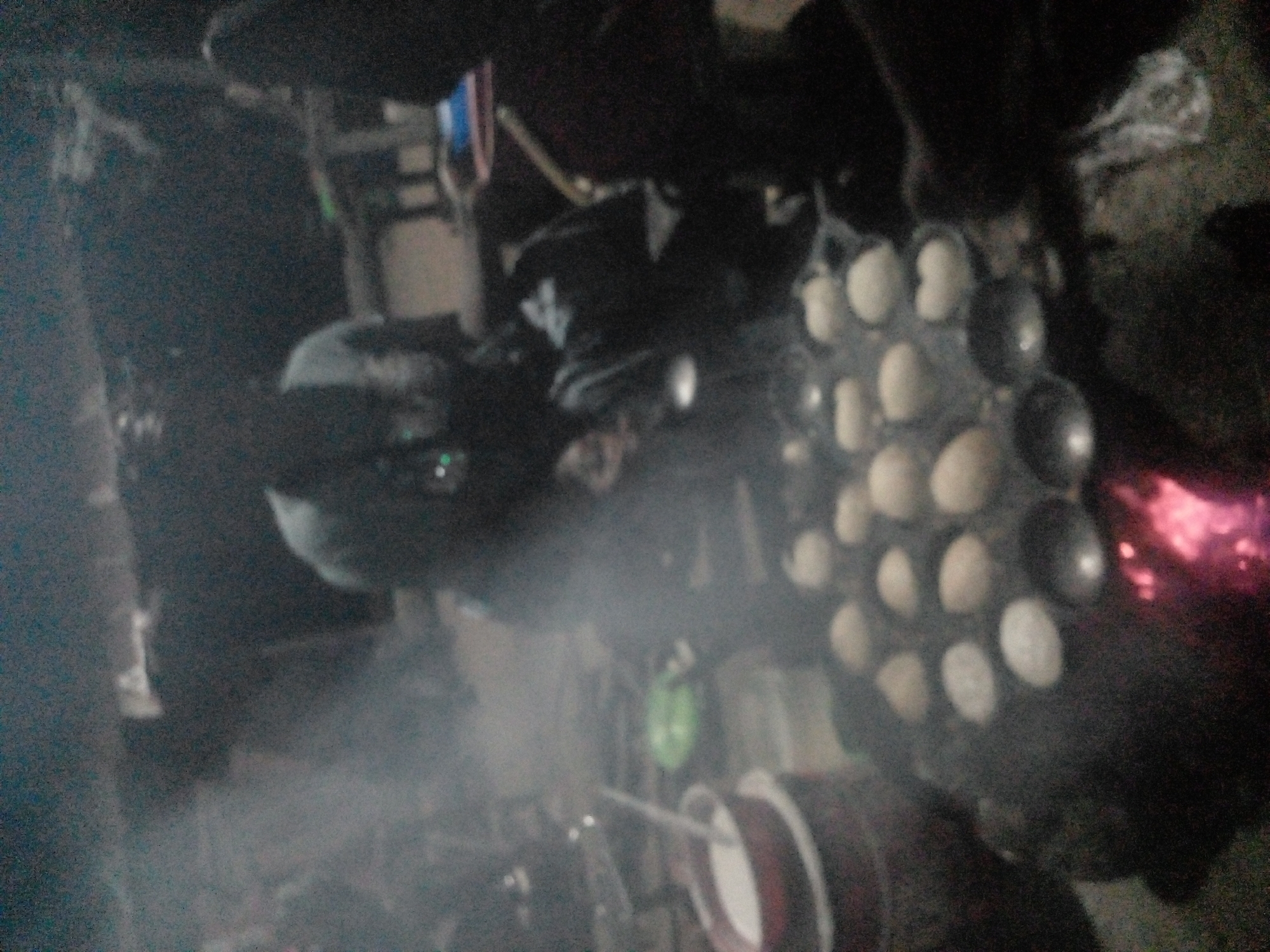 In the city of Langtang, Nigeria, this image captures a dimly lit outdoor scene, likely at night, focusing on a cooking area. A person, mostly obscured by steam and shadow, is actively engaged in preparing food on a flat cooking surface with multiple circular indentations, resembling a specialized pan for small, round cakes or dumplings. This cooking apparatus is positioned over what appears to be a heat source, indicated by a faint red glow and flickering light at the bottom of the frame, suggesting embers or a low fire.
To the left, a dark red bowl containing a white powdery substance, possibly flour or batter, is visible. A utensil, such as a spoon or whisk, rests inside the bowl, hinting at the preparation of the food being cooked. Several of these round food items are already cooked and rest in their respective indentations on the pan, appearing light in color. Steam rises from the cooking area, obscuring parts of the scene and adding to the atmospheric effect.
The background is dark and indistinct, with blurry shapes suggesting a rustic or informal outdoor setting. The overall impression is one of food preparation in a humble, possibly communal, environment, characterized by low light conditions and the visible process of cooking.