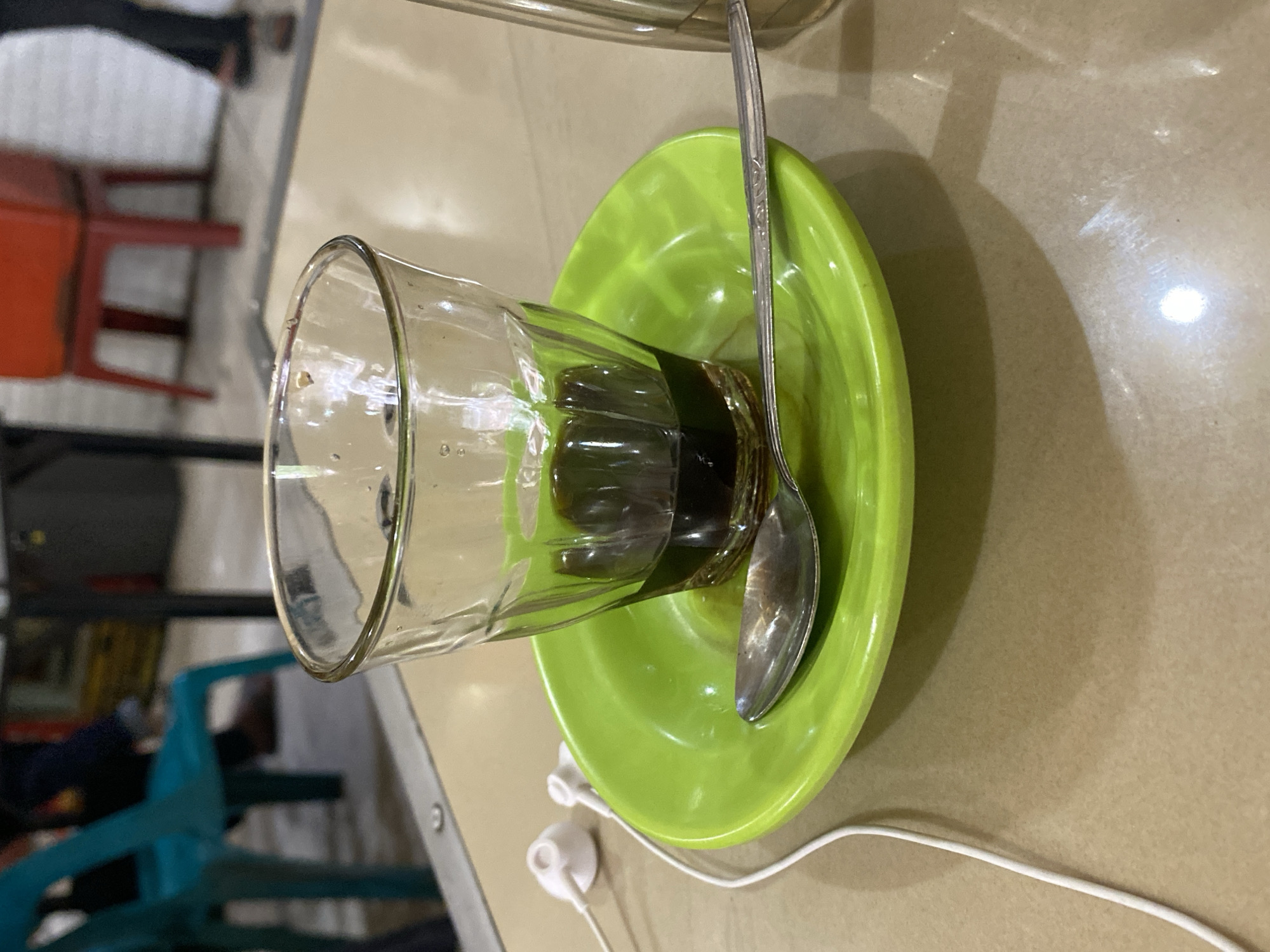 A close-up, eye-level shot focuses on a glass of dark liquid, likely coffee, sitting on a bright green saucer. A silver spoon rests on the saucer. To the left of the saucer, a pair of white earbuds and their white cord are visible on a light brown table. In the background, out of focus, are parts of chairs and people, suggesting an indoor setting like a cafe or restaurant. The lighting appears to be from above, creating a soft reflection on the table surface. The image does not provide specific visual cues to pinpoint its location within Lhokseumawe, Indonesia, beyond inferring a public establishment.