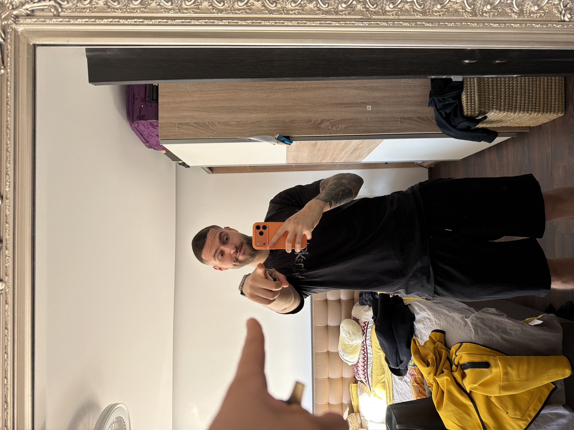 A man stands in front of a mirror, holding an orange mobile phone and pointing towards the camera with one hand, while the other hand makes a thumbs-up gesture. He is wearing a black t-shirt and black shorts. The mirror reflects a room with a wardrobe in the background, part of a bed with various clothes and pillows, and a wall. A fan is also visible on the left side of the reflection. The location of the image is likely inside a residential space, but information to determine it is in Sofia, Bulgaria, is not available.