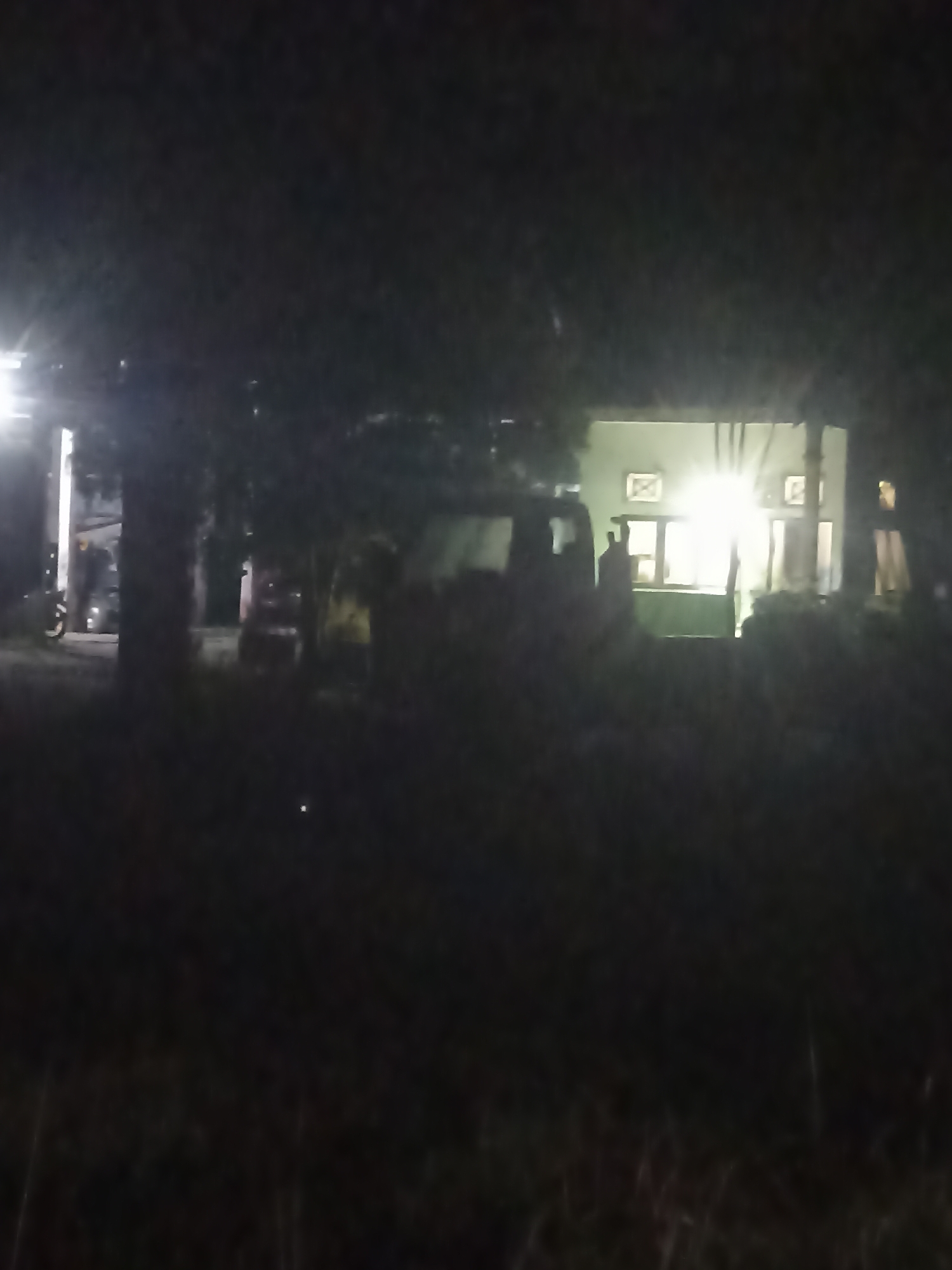 The image depicts a dark, outdoor scene at night, possibly in Kampung Tengah, Indonesia. A brightly lit building with windows is visible in the background, suggesting it's illuminated from within. In front of the building, there's a dark-colored van or large vehicle. To the left, a motorcycle is parked under a light source. Trees and vegetation are present, adding to the outdoor setting. The overall impression is of a nighttime scene with some artificial lighting.