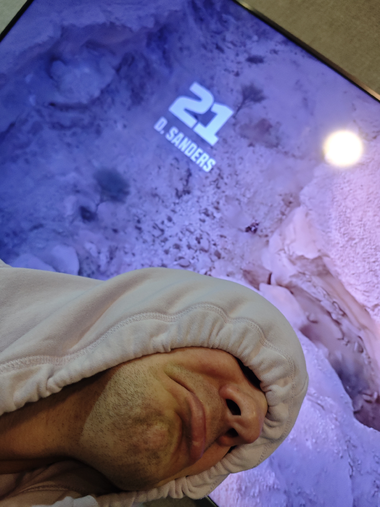 A person is seen with part of their face visible, covered in the hood of a light-colored sweatshirt. In the background, a television screen displays the number 21 and the name D. SANDERS. The background of the television shows a landscape. The location is not discernible as Chaikovsky, Russia based on the given image.