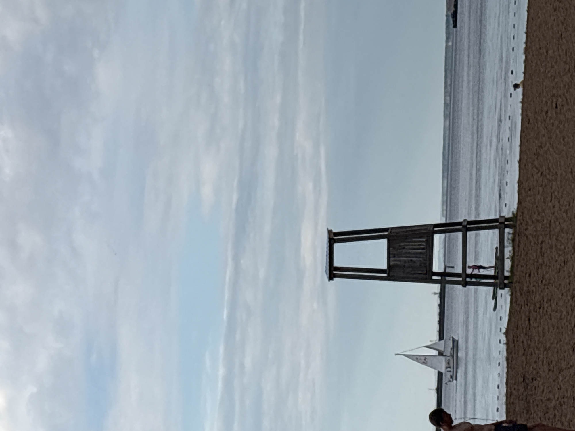 A wooden lifeguard tower stands on a sandy beach beside a calm body of water. The sky is cloudy with streaks of light blue. A sailboat with white sails navigates the water in the distance. A person is visible on the beach near the lifeguard tower, appearing to be sunbathing or resting. The image is oriented vertically.