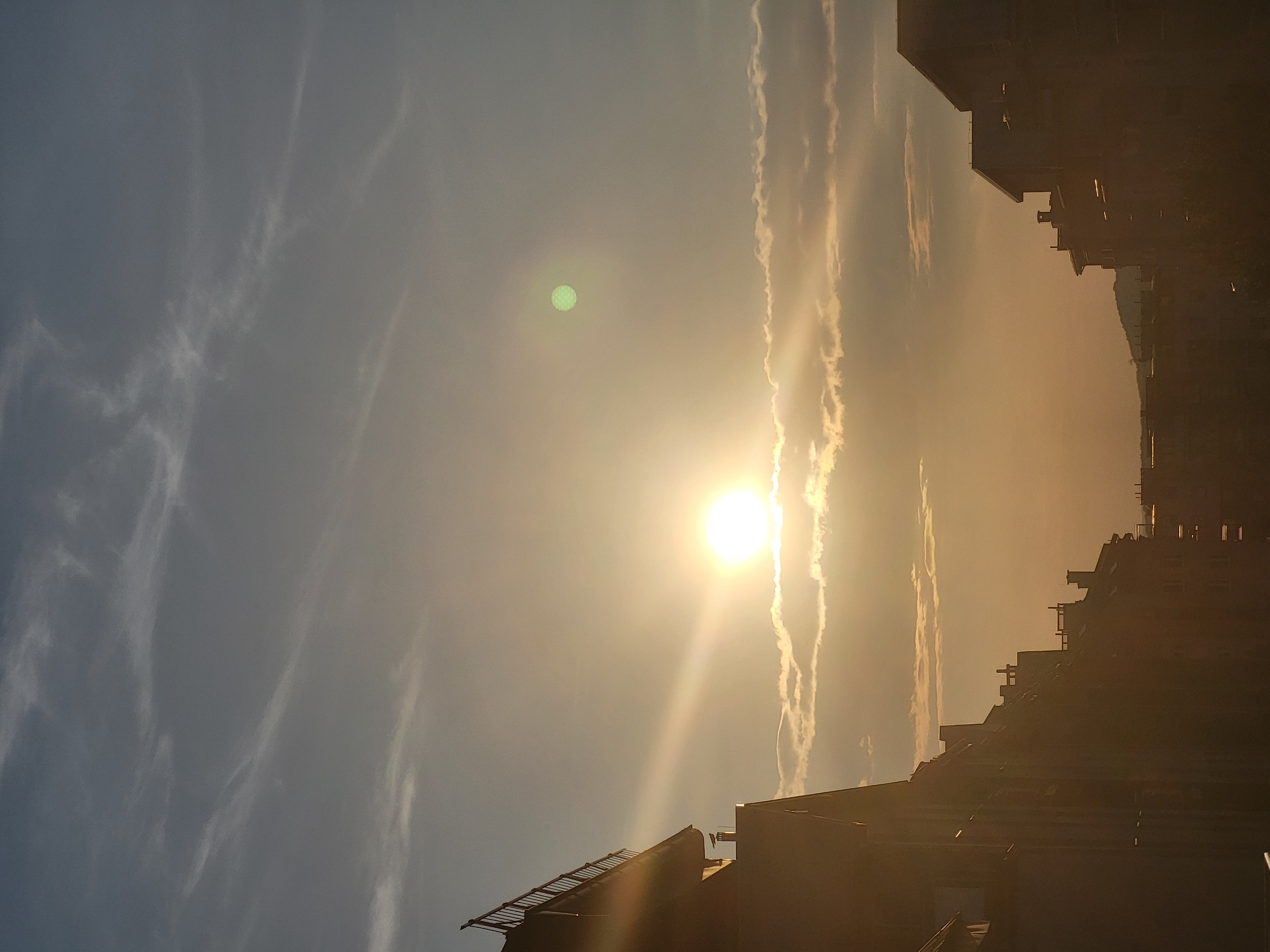 The image depicts a city skyline at sunset. The sun, a bright orb, is positioned in the center of the frame, casting a warm glow and long rays of light across the sky. Wispy clouds are scattered across the horizon, some illuminated by the setting sun. Silhouetted buildings with visible rooftops and chimneys frame the lower portion of the image, with one building on the left having a railing along its edge. A faint green circular lens flare is visible in the upper right quadrant of the sky. The overall scene suggests a peaceful, late afternoon or early evening in an urban environment. The location is Unknown, Unknown.