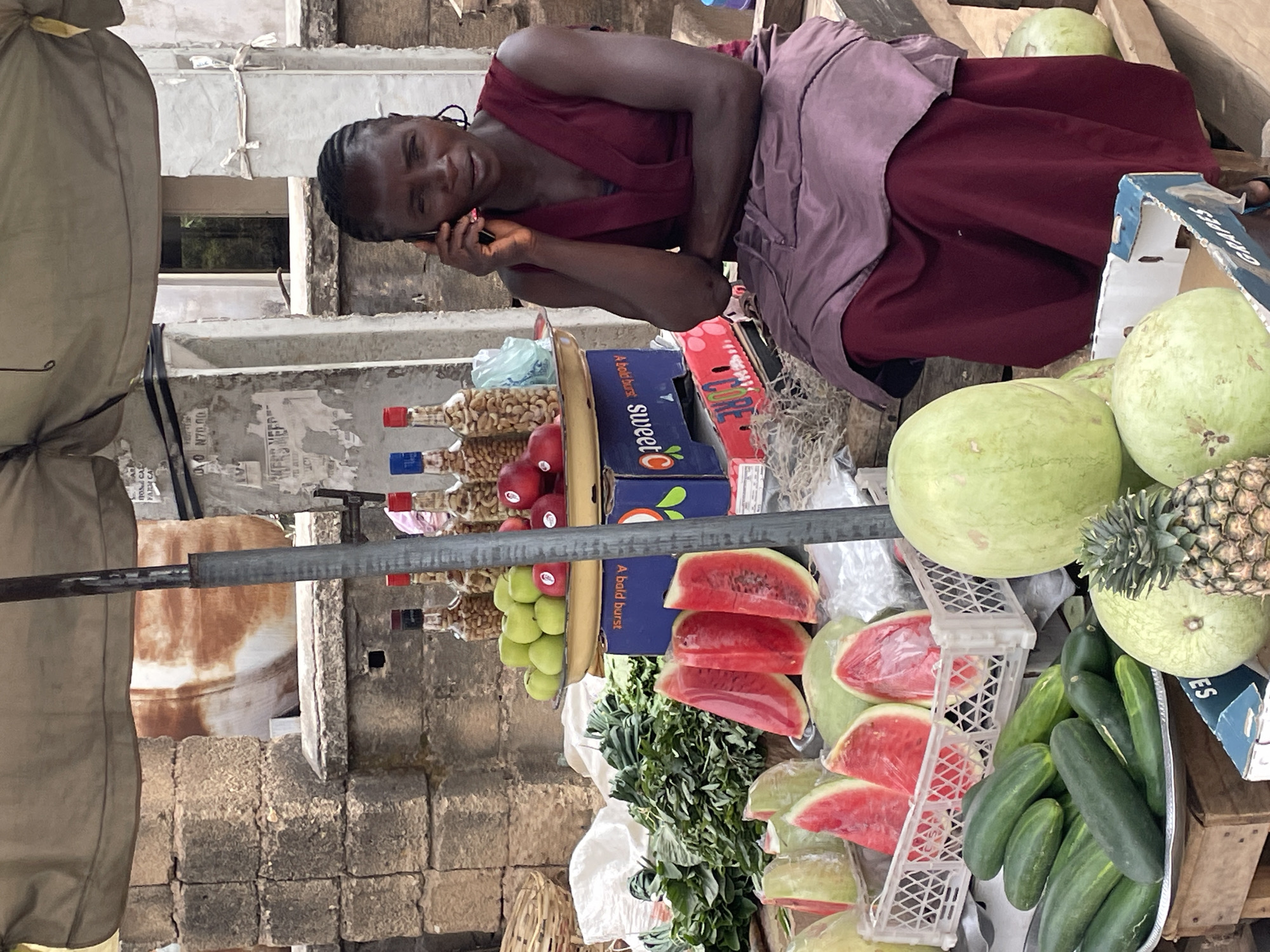 An outdoor market scene features a woman vendor at her produce stall, captured during daylight hours. The central figure is an adult woman with dark skin and braided hair, wearing a dark red top and a light purple apron. She is actively engaged in a phone conversation, holding the device to her right ear, with a subtle, relaxed smile on her face.
Her stall is abundantly stocked with fresh produce. Prominently displayed are several whole green watermelons, alongside numerous slices of watermelon, some individually wrapped in clear plastic. A pineapple with its crown is visible near a whole watermelon. Long green cucumbers are neatly arranged in a metal tray, while bunches of leafy green vegetables are also part of the display. Other fruits include green apples and what appear to be red apples, as well as several whole, light green gourds or squash. Packaged goods like bottles filled with nuts (possibly cashews and peanuts) are stacked on a shelf.
The produce is housed in various containers, including cardboard boxes with visible branding such as "sweetC," "CORE," "GRAPES," and "A bold burst." White plastic crates also hold some of the sliced watermelon. A sturdy metal pole, likely part of the stall's structure, is visible in the foreground.
The background reveals weathered concrete block walls and plaster, indicating an urban or semi-urban setting consistent with Jos, Nigeria. A window and parts of large, light brown sacks are also visible. Faint text, including "N70.00," can be seen on one of the background walls. The scene depicts a lively market environment with fresh goods on offer.