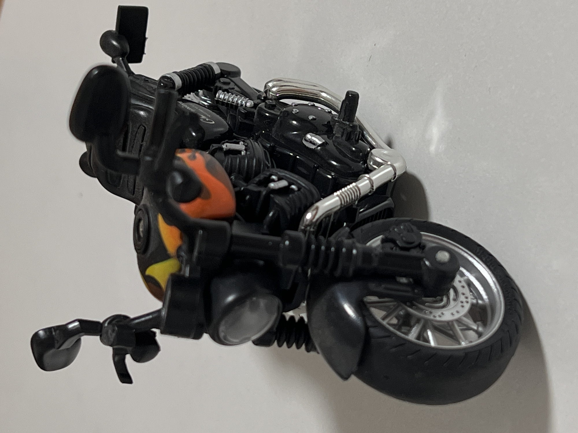 The image shows a close-up, high-angle view of a black toy motorcycle with orange and yellow flame decals on the fuel tank. The motorcycle is positioned on a white surface, likely a tabletop or floor. Details visible include the front wheel with a silver rim and black tire, a silver brake disc, the front suspension with black bellows, a round headlight, handlebars with black grips and mirrors, and parts of the engine and exhaust system. The motorcycle is not in motion. There are no people or other objects visible in the immediate vicinity. The background is a plain white wall. The image does not contain any information that would allow for its location to be determined as Qarağandı city administration, Kazakhstan.