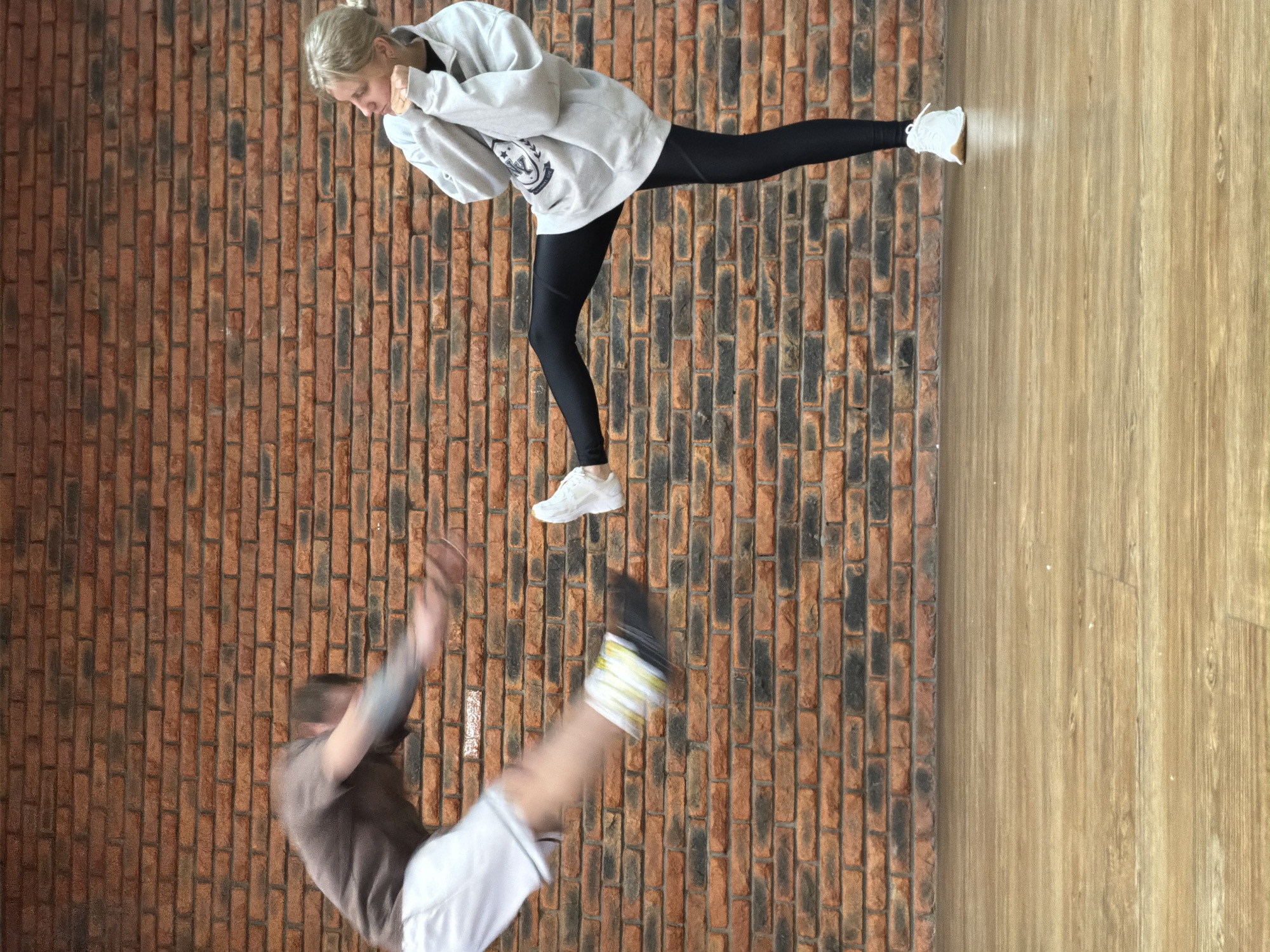 A woman with blonde hair tied in a bun, wearing a white hooded sweatshirt, black leggings, and white sneakers, is depicted in a martial arts pose. Her right leg is extended forward, and her hands are near her face in a defensive or offensive stance. Below and slightly to the left, a man in a brown shirt and white shorts is captured mid-air in a dynamic pose, with one leg extended upwards. The background consists of a brick wall on the left and a wood-look floor on the right. The orientation of the image appears to be rotated 90 degrees counterclockwise. The location is Chaikovsky, Russia.