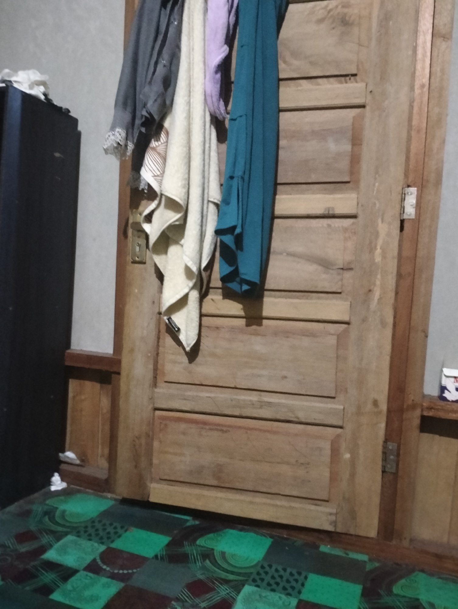 The image depicts an interior scene, likely a doorway, in a dimly lit room. A wooden door, segmented into several panels, dominates the right side of the frame. Several pieces of clothing, including a gray shirt, a beige towel, and a teal garment, are hanging from an unseen hook or rail to the left of the door. To the far left, a dark, rectangular object, possibly a cabinet or dresser, is partially visible, with some white material resting on top. The floor is covered with a patterned mat, featuring green, maroon, and black squares. The overall impression is one of a utilitarian living space. The location is identified as Mandala Jaya, Indonesia, suggesting a domestic setting within that region. There are no people or animals present in the image.