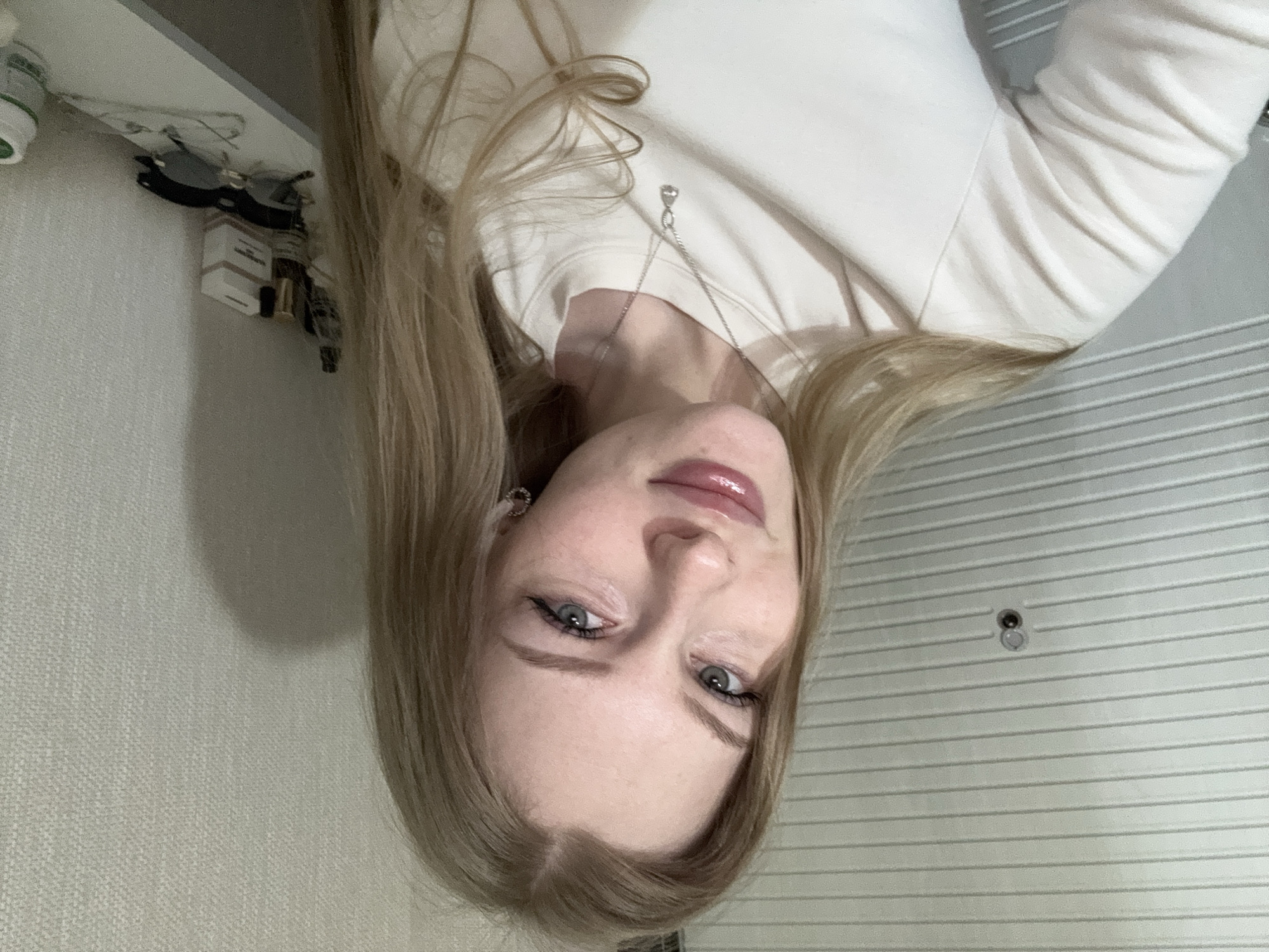 The image is a close-up, inverted selfie of a young woman with blonde hair. She is wearing a light-colored, long-sleeved shirt and a silver necklace. Her features include fair skin, blue eyes, and glossy lips. An earring is visible in her left ear. The background is a wall with horizontal lines and a peephole. In the upper-left corner, there's a shelf with various items, including bottles and packaged goods. The location, Perm, Russia, cannot be determined from this image.