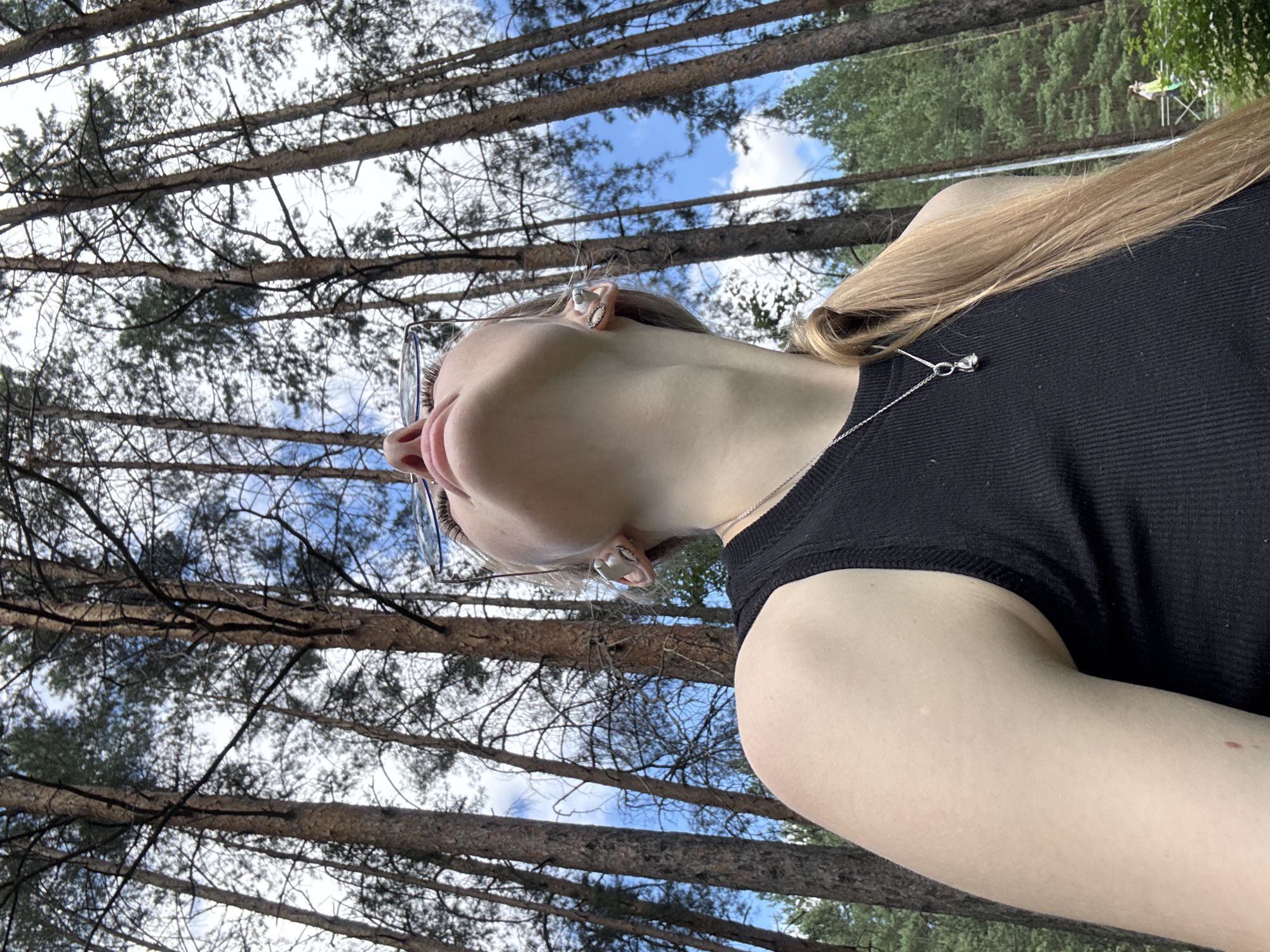 The image shows a young woman looking up in a forest. She has long blonde hair pulled back in a ponytail and is wearing a black tank top and a silver necklace. She is also wearing glasses and has on large, decorative earrings. The trees in the background are tall and thin, with sparse foliage, suggesting it may be autumn or early spring. The sky is blue with some white clouds. There is a hint of a wooden structure in the background on the right, and a person walking in the distance. The exact location is not identifiable but the environment appears to be a natural, wooded area.