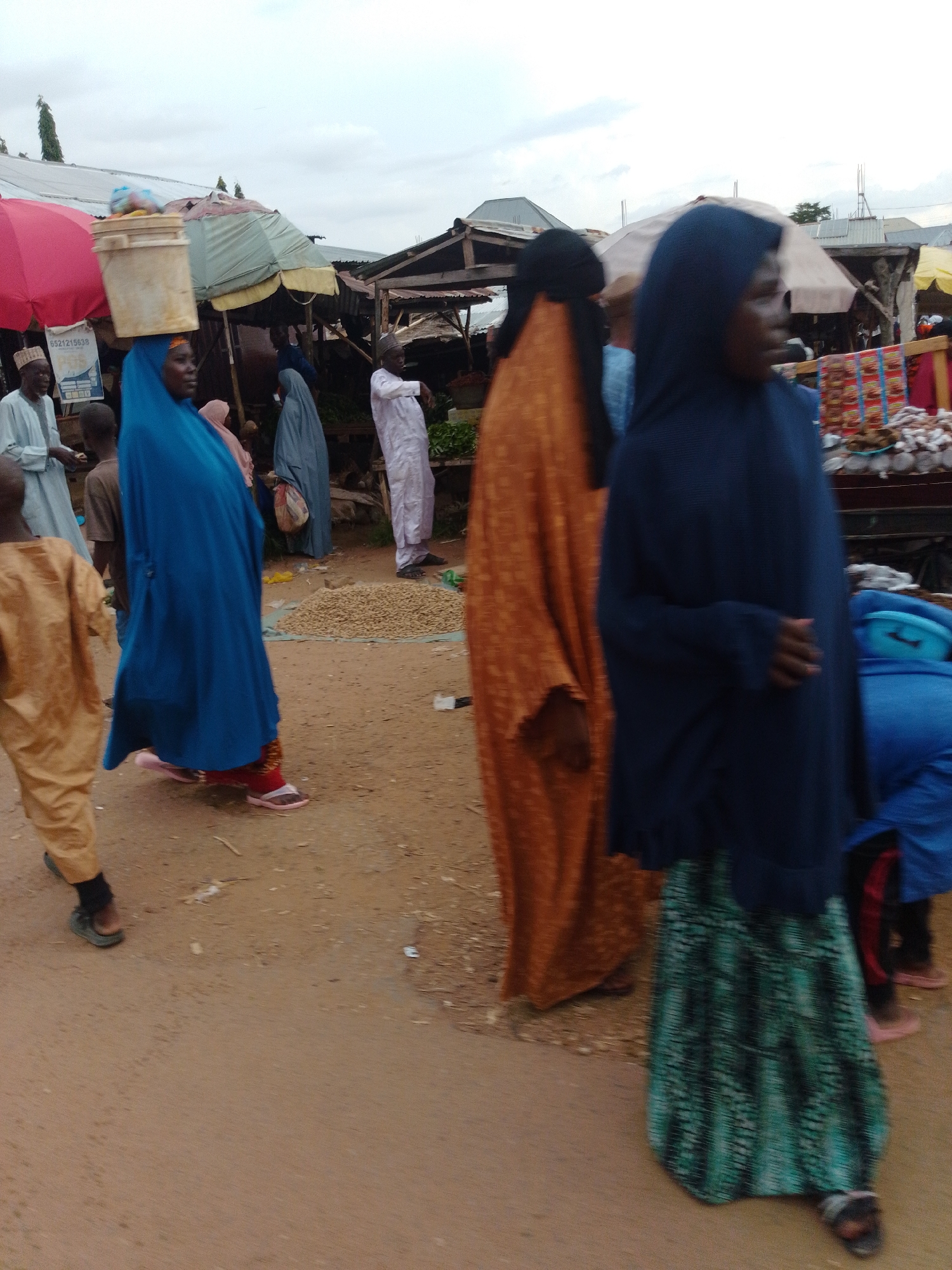 A market scene in Gombe, Nigeria, shows several individuals in traditional clothing amidst stalls and produce. A woman in a blue hijab and a beige bucket on her head walks past a stall. To her left, a person in a yellow garment stands. Behind them, a woman in an orange abaya is visible, and further back, a person in a blue cloak walks away. The ground is dirt, with a pile of nuts or seeds in the background. The sky is overcast, suggesting a cloudy day. The overall atmosphere is that of a busy marketplace.