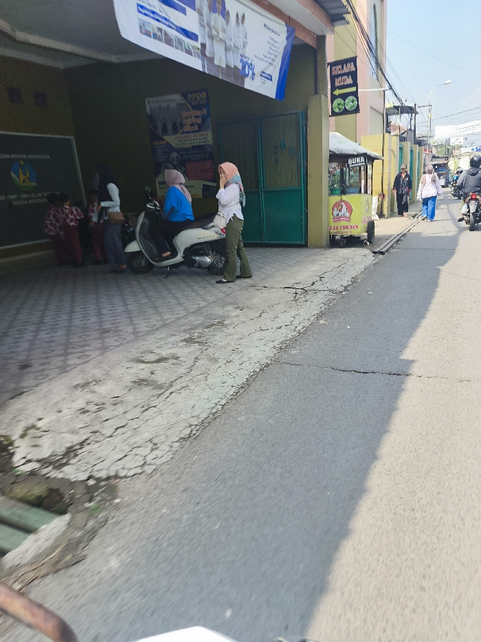 The image shows a street scene in Cileunyi, Indonesia (latitude: -6.941, longitude: 107.726). A white moped is parked on the sidewalk, while a group of people stand around, some talking and some looking at the moped. One woman in a blue shirt sits on the moped, her legs dangling. A woman in a white shirt and headscarf stands nearby. A small yellow food cart with the sign “BAH CHICKEN” is parked near the sidewalk on the right side of the image. A green gate is behind the food cart. A sign with the words “KELAPA MUDA” is visible on the wall of a building. The road is paved and shows signs of cracking. The sidewalk is also paved. There are several other people walking on the street in the distance. The sky is clear and blue. The trees in the background are bare.