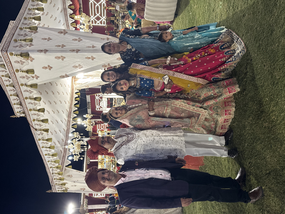 The image captures a joyful gathering of people, likely an Indian celebration such as a wedding or engagement, held outdoors in the evening or at night. The dark sky is visible at the top left, illuminated by various decorative lights and chandeliers hanging from elaborate white canopies or tents that cover the area. The ground beneath the attendees is green grass.

In the foreground, a group of individuals, primarily adults and one small child, are posing for a photograph. They are all dressed in festive, traditional Indian attire.

From left to right in the foreground:
*   A man stands wearing a brown turban, a light pink collared shirt, and a dark jacket over dark trousers. He is looking towards the camera with a gentle smile.
*   Next to him, an older man is dressed in a white kurta pajama set, a traditional red turban, and glasses. He is also smiling at the camera.
*   Behind these two men, a woman is seen wearing a heavily embroidered light pink and gold lehenga with a matching dupatta. She has bangles on her wrists and is smiling brightly.
*   Next to her, another woman is dressed in a vibrant outfit consisting of a blue top, a bright yellow dupatta draped over her shoulders, and a richly embroidered red lehenga. She is holding a white smartphone in her hand and smiling at the camera.
*   Slightly behind and to the right, another woman wears a dark colored top and a red lehenga with a patterned dupatta. She is smiling.
*   Further to the right, a woman in a dark top and a light blue or turquoise colored outfit holds a small child, possibly a baby or toddler, who is also dressed in a light blue garment. Both the woman and the child are looking towards the camera, and the woman is smiling.

The background features more decorative elements, including an illuminated latticework structure and additional lighting, confirming the celebratory atmosphere. All visible faces in the main group show happy and smiling expressions, indicating a joyous occasion. No discernible text is visible in the image.