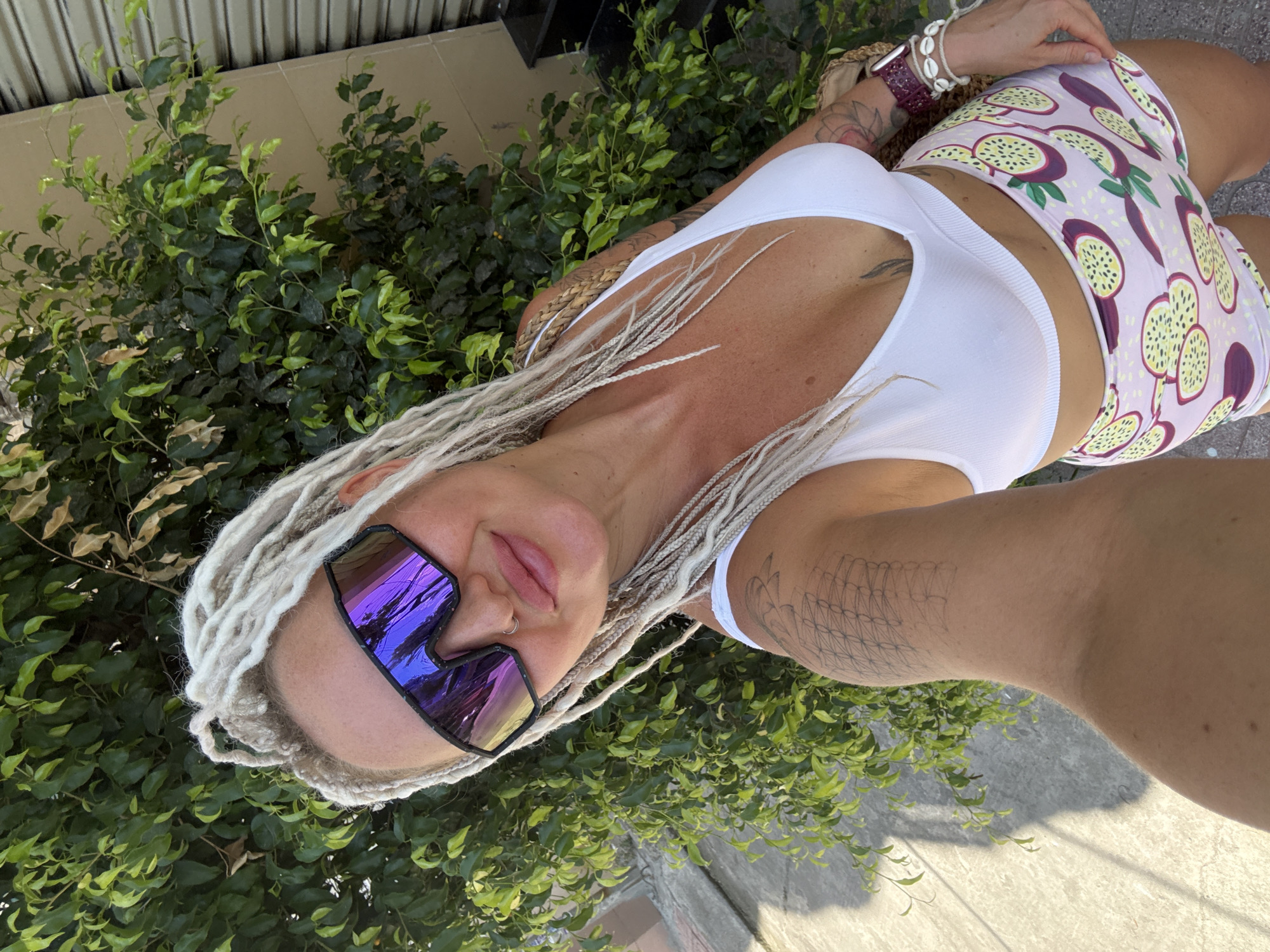 A woman with blonde dreadlocks and large, reflective sunglasses is taking a selfie outdoors. She is wearing a white crop top and patterned shorts. Behind her, a lush green bush and a slatted wall are visible. The scene appears to be a sunny day. The location is identified as Bac Nha Trang Ward, Vietnam.
