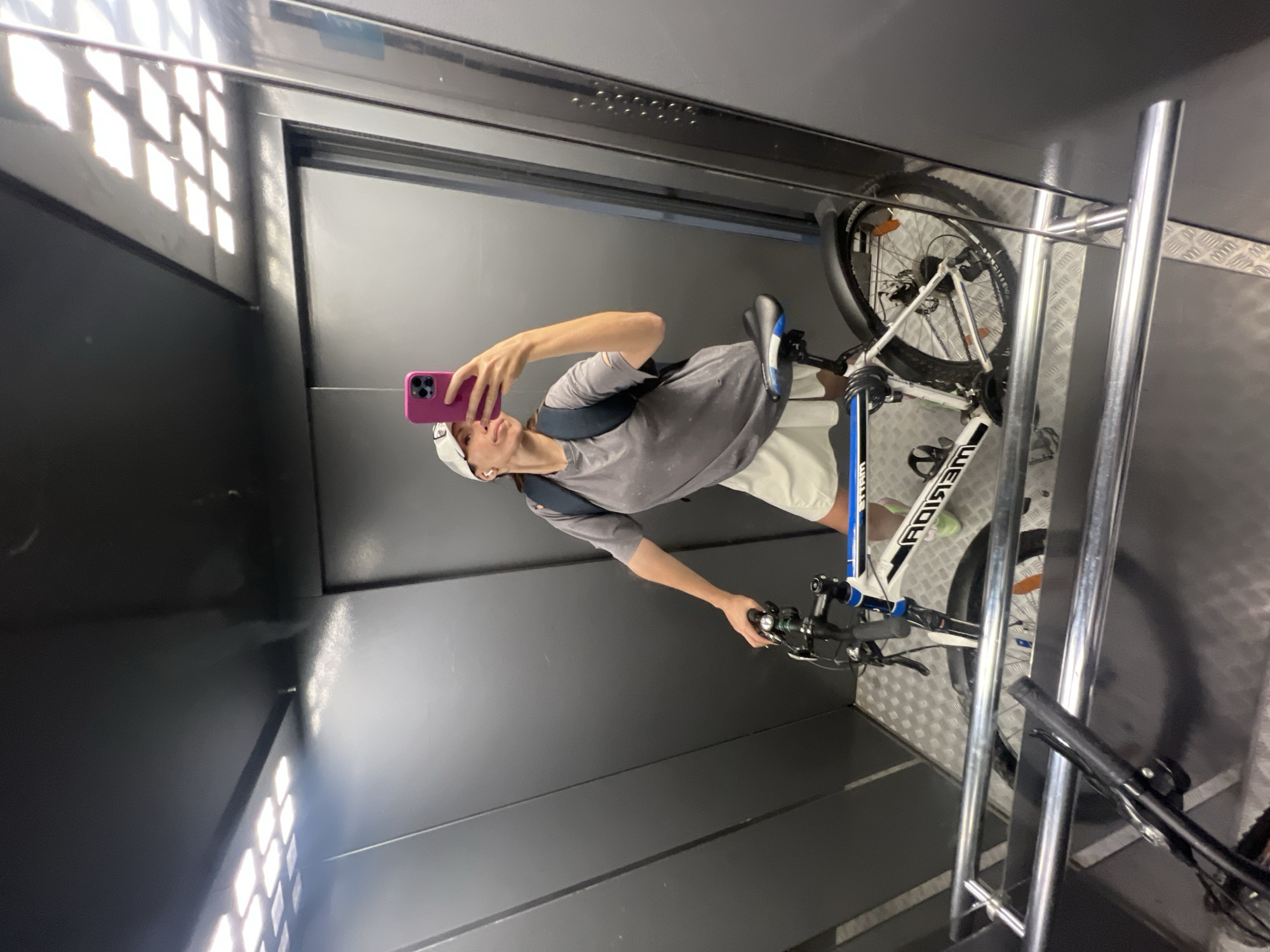 A person stands in an elevator, holding a pink-cased phone and taking a selfie. Next to them is a white and blue mountain bike, leaning against the elevator wall and bike rack. The elevator interior has gray walls, diamond-patterned flooring, and grid-patterned lighting. The scene appears to be indoors, possibly in a residential or commercial building with elevator access. There is no definitive indication of a specific city like Moscow, Russia.