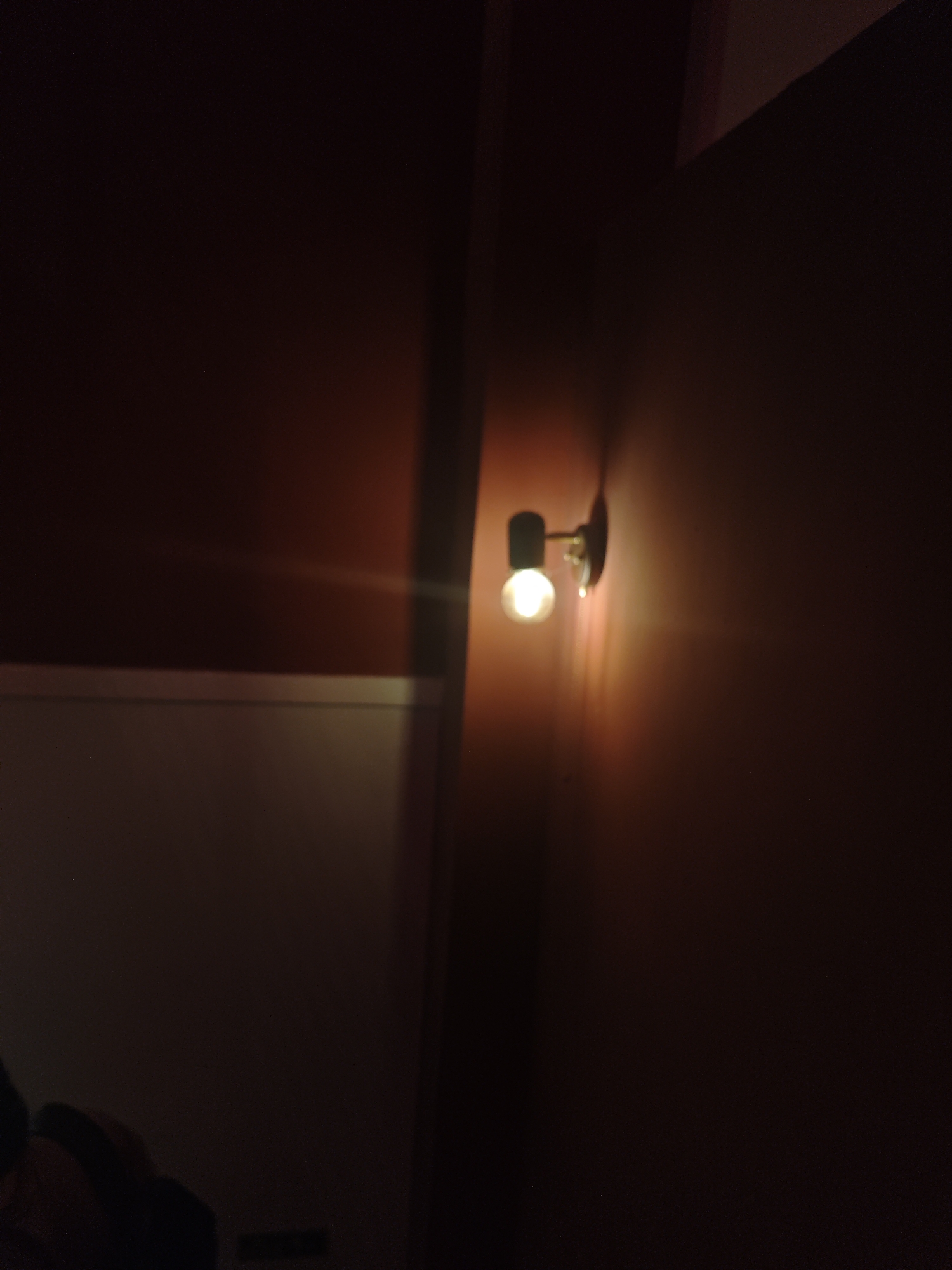 A dim interior scene is dominated by the warm glow of a wall-mounted light fixture. The fixture is a cylindrical shape with a gold or brass mounting. The light illuminates a brown wall and a partially visible white wall to the left. Shadows are prominent throughout, obscuring details in the lower portion of the image. The location and specific subjects are not identifiable from the image. There is no way to confirm this image is in Chongqing High-tech Zone, China.