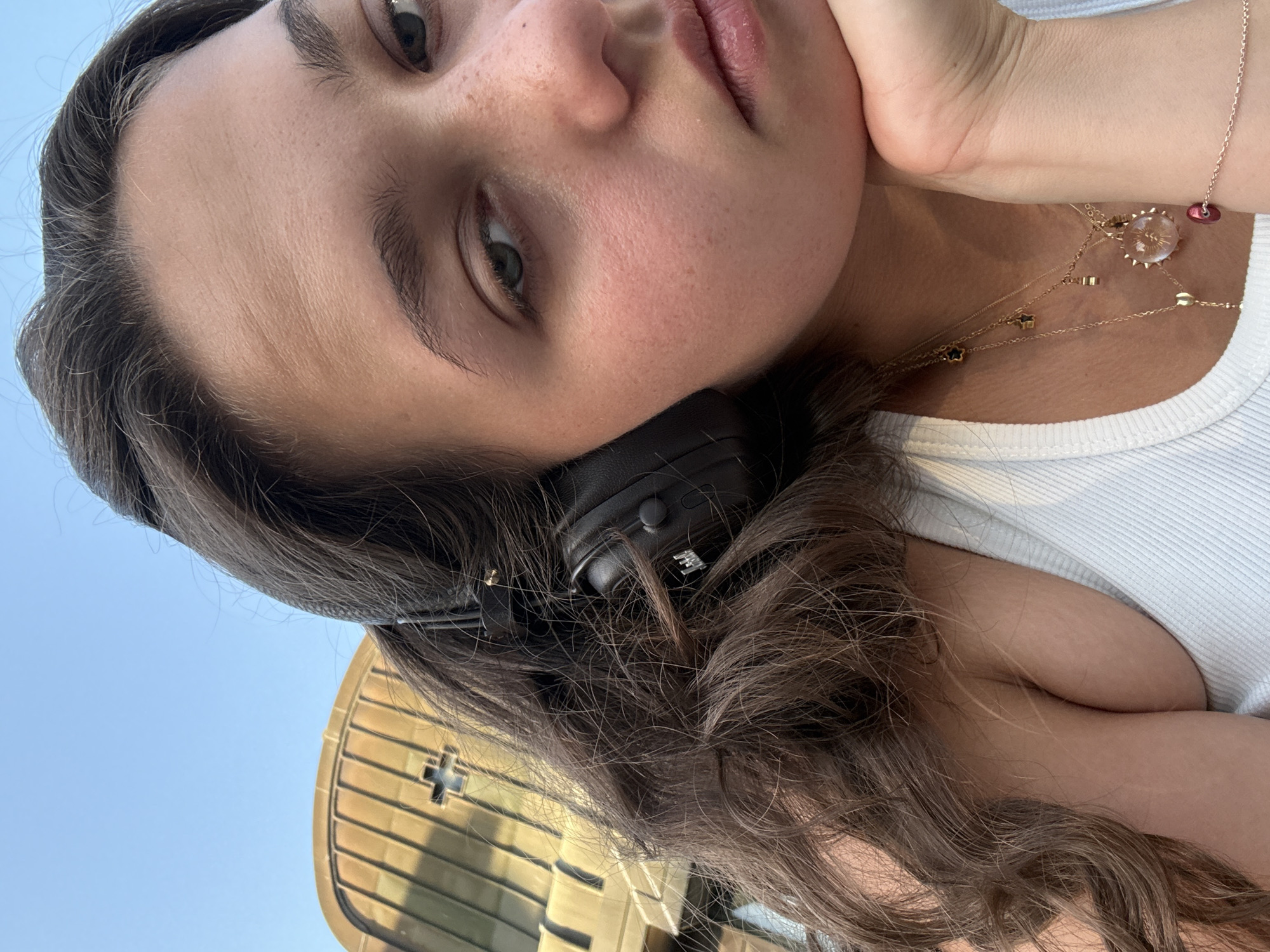A close-up, low-angle selfie shows a young woman with long, dark, wavy hair wearing black headphones and a white tank top. She rests her chin on her hand, and her gaze is directed towards the camera with a neutral expression. Multiple gold necklaces are visible around her neck, one with a pendant featuring a sun-like design. In the background, a curved, tan-colored building with vertical lines and a cross-shaped window is visible against a clear blue sky. The woman's skin has a light complexion with visible freckles. The overall scene appears to be outdoors during the day. The architecture suggests a possible religious or historical building, and given the style, it could be located in Milan, Italy, possibly near a church or monument with such features.