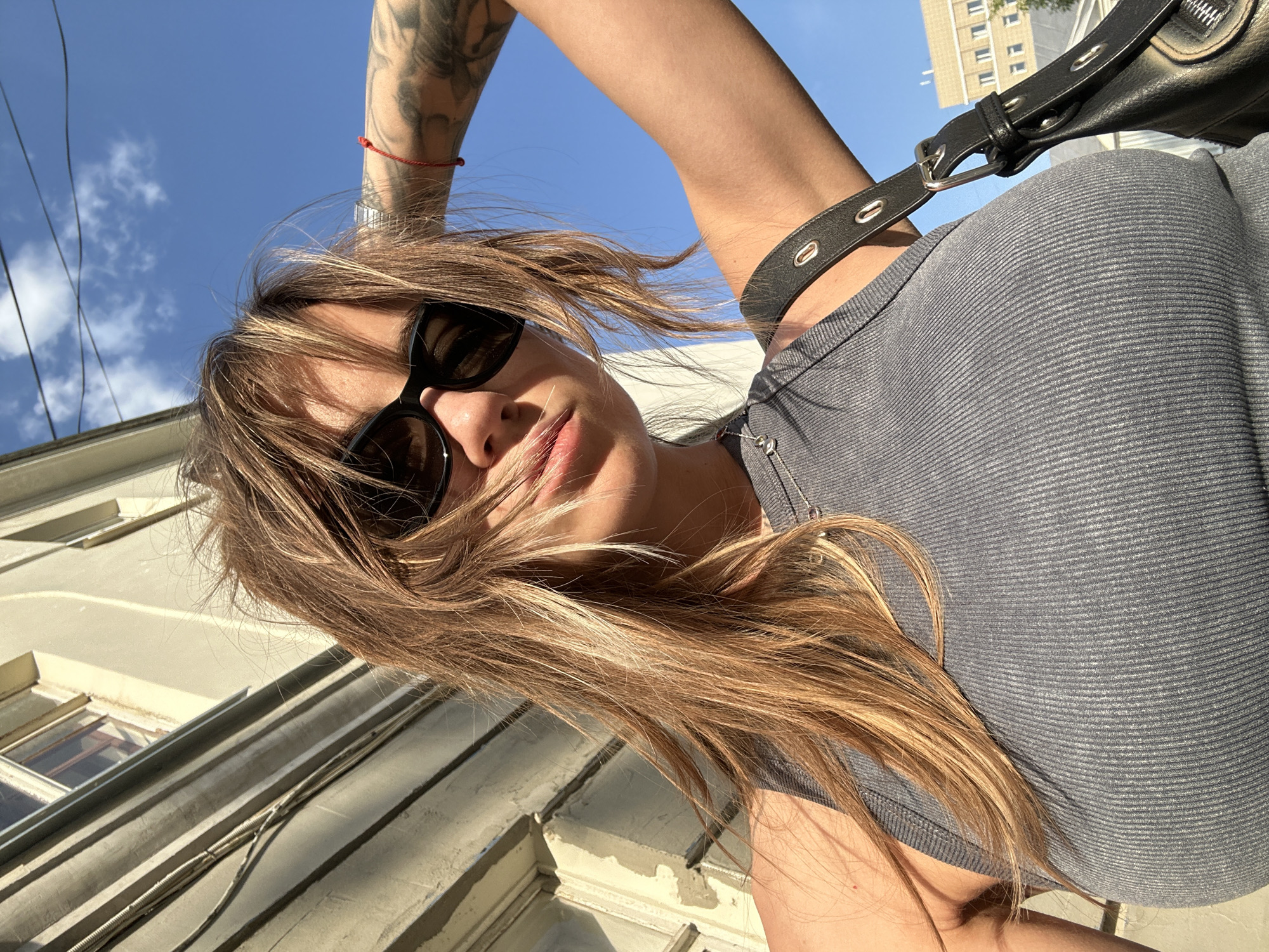 A low-angle perspective selfie captures a woman with shoulder-length brown hair, blonde highlights, and sunglasses, standing in Moscow, Russia. She wears a gray ribbed tank top and a black shoulder bag with a leather strap. A tattoo is visible on her raised arm, and she has a red string bracelet. The background features a clear blue sky and part of a light-colored building with windows.