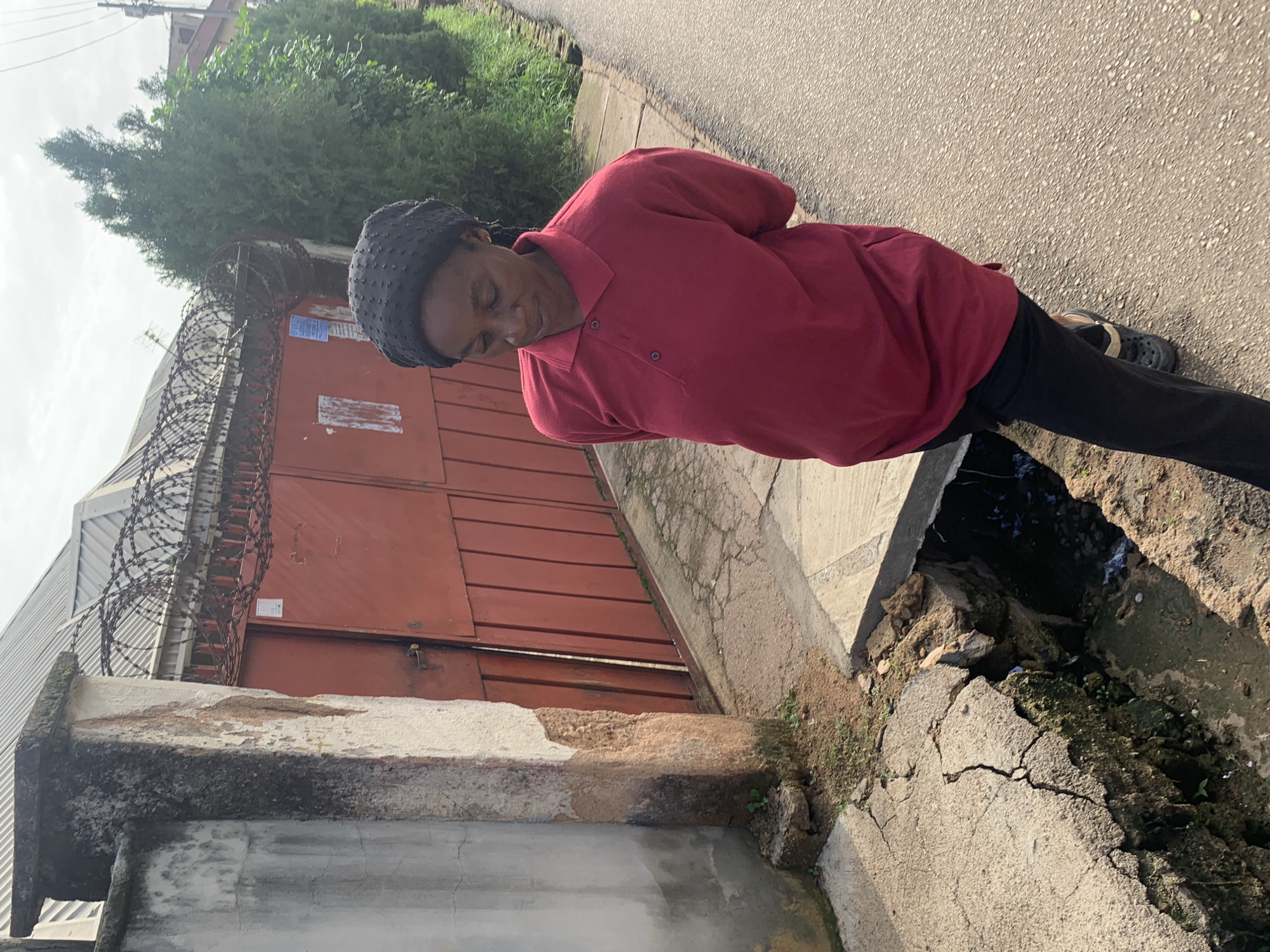 In Jos, Nigeria, a woman with dark skin and braided hair is walking on a paved road. She is wearing a dark headscarf and a maroon polo shirt, with her hands clasped behind her back. To her left, a red metal gate topped with barbed wire is visible, and beside it, a concrete wall shows signs of damage and weathering. A large, open drain with cracked concrete edges is situated in front of the woman, revealing murky water and debris. The background features a row of tall, green cypress trees and a building with a corrugated metal roof. The overall scene depicts an urban environment with elements of disrepair.