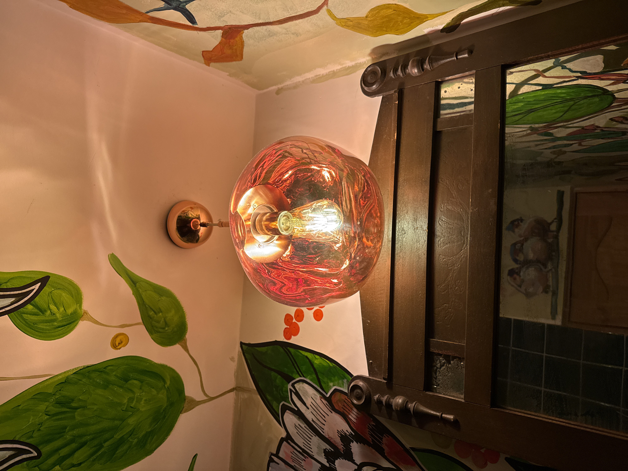 The image shows an interior scene, presumably a bathroom, in Yerevan, Armenia. The walls are painted with a mural of green leaves, white and pink flowers, and small red dots. A round, orange glass light fixture with an exposed light bulb is mounted on the wall. A dark brown wooden cabinet or mirror frame is positioned next to the light fixture, reflecting the mural on the walls and ceiling. The ceiling is also painted with images of leaves and other objects. The scene appears to be lit by the light fixture itself, creating warm tones.
