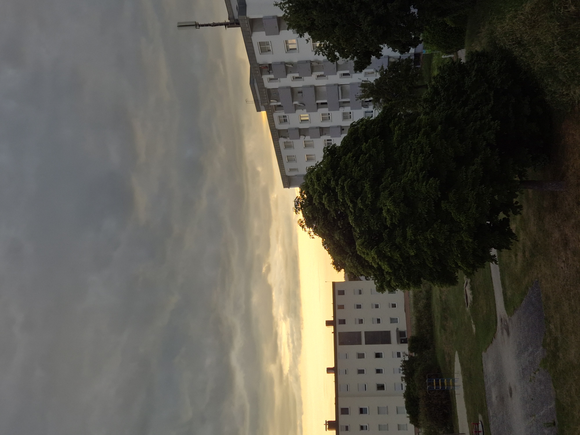 The image depicts an urban landscape at either sunrise or sunset, characterized by a heavily overcast sky with dark grey clouds overhead transitioning to a bright yellow-orange glow at the horizon. Two multi-story residential buildings dominate the scene.
On the left, a light-colored apartment building with a flat roof features multiple rows of rectangular windows, many equipped with white roller shutters. Darker vertical panels are present on its facade.
On the right, another similar apartment building, predominantly white, exhibits multiple balconies with light grey railings and numerous windows. Several satellite dishes are affixed to the balconies. A tall, slender cellular antenna or telecommunications mast is mounted on the roof of this building, extending into the sky on the far right.
A large, dense, dark green deciduous tree stands prominently in the mid-ground, partially obscuring the right building. Additional trees and shrubs are scattered throughout the scene.
The foreground and mid-ground feature a grassy area, a paved section with dark, irregular patches, and a light-colored patch resembling a sandpit. A blue and yellow metal climbing frame, consistent with playground equipment, is visible in the grassy area towards the left side of the frame. No individuals are present in the visible area.